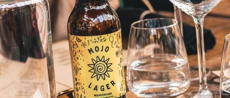 Una botella de cerveza Mojo Lager con etiqueta que muestra un sol y varios iconos, rodeada de vasos vacíos sobre una mesa de madera.