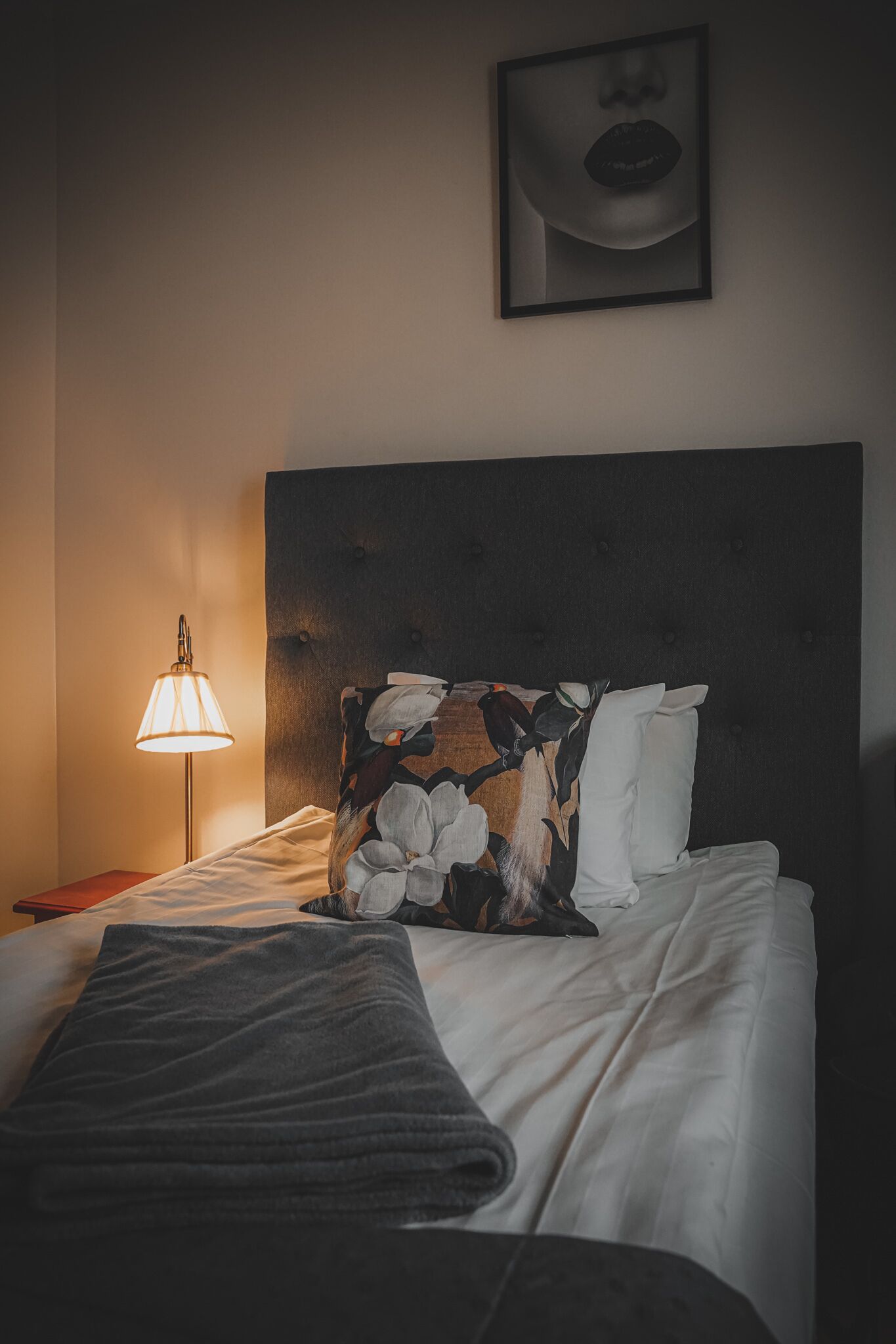 Una cama bien hecha con un cabecero oscuro, una almohada estampada con motivos florales y una almohada blanca. Una lámpara con pantalla blanca está sobre la mesita de noche junto a la cama.
