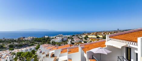 A coastal residential area with terracotta-tiled rooftops, white buildings, and a clear view of the sea and distant mountains.