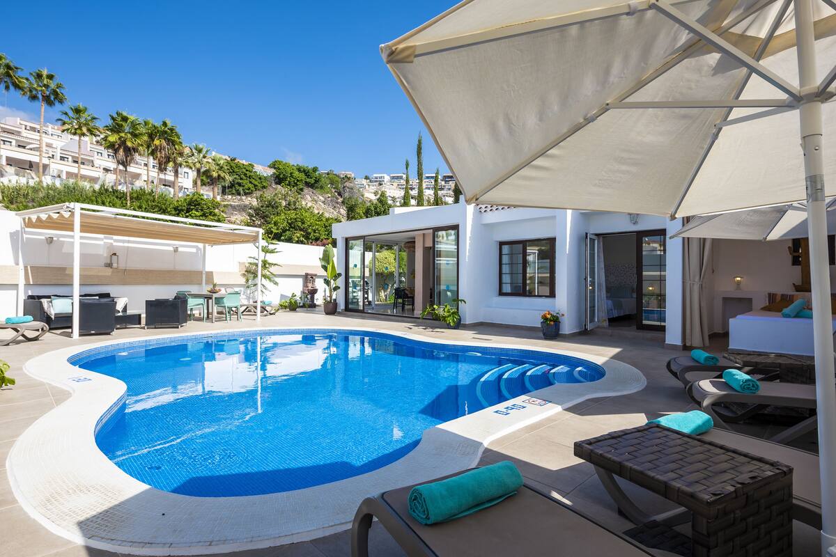 Una piscina con sillones de descanso y sombrillas, rodeada por un edificio con puertas de vidrio y vistas a palmeras.