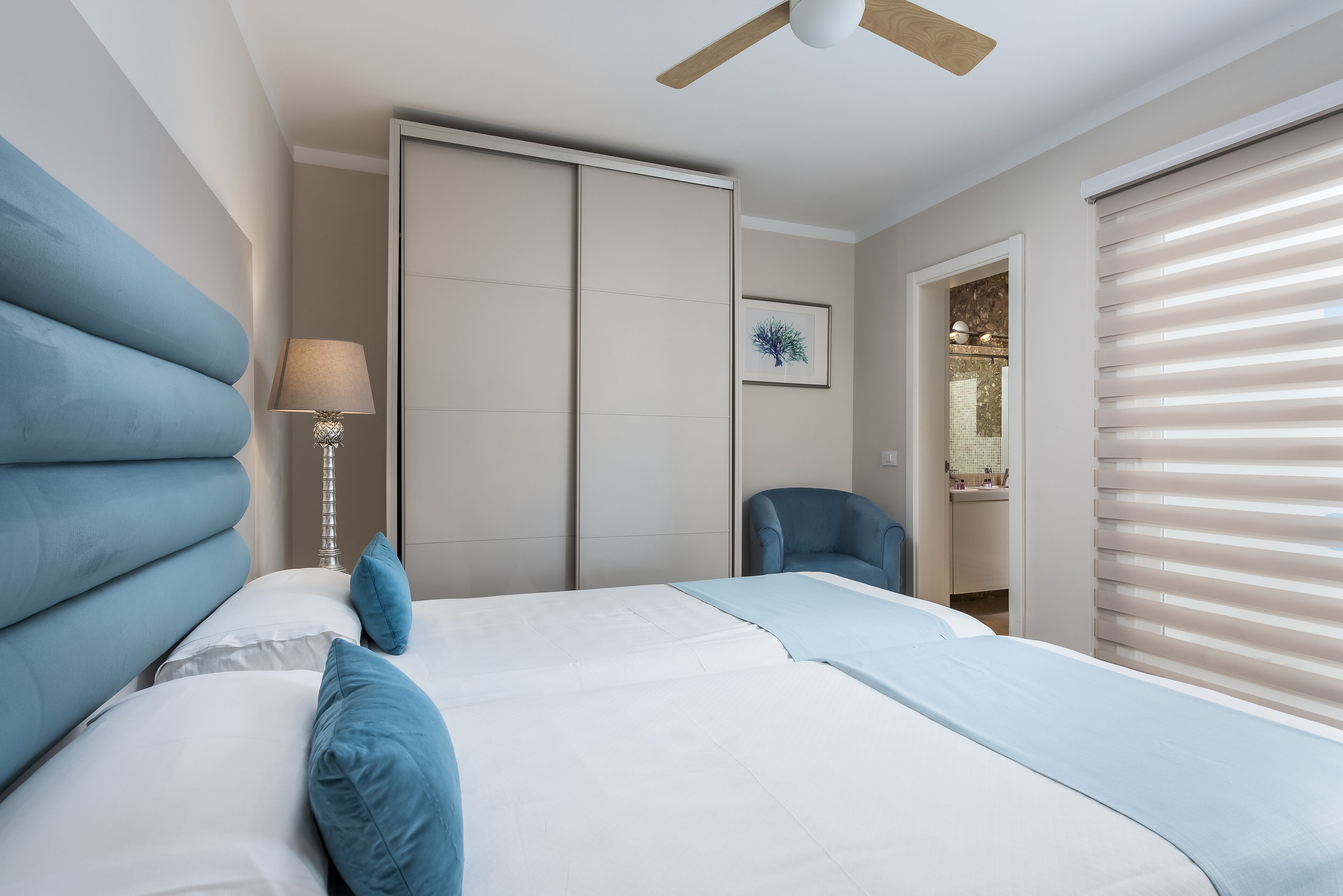A bedroom with a bed, a chair, a lamp, a ceiling fan, and a window with blinds.