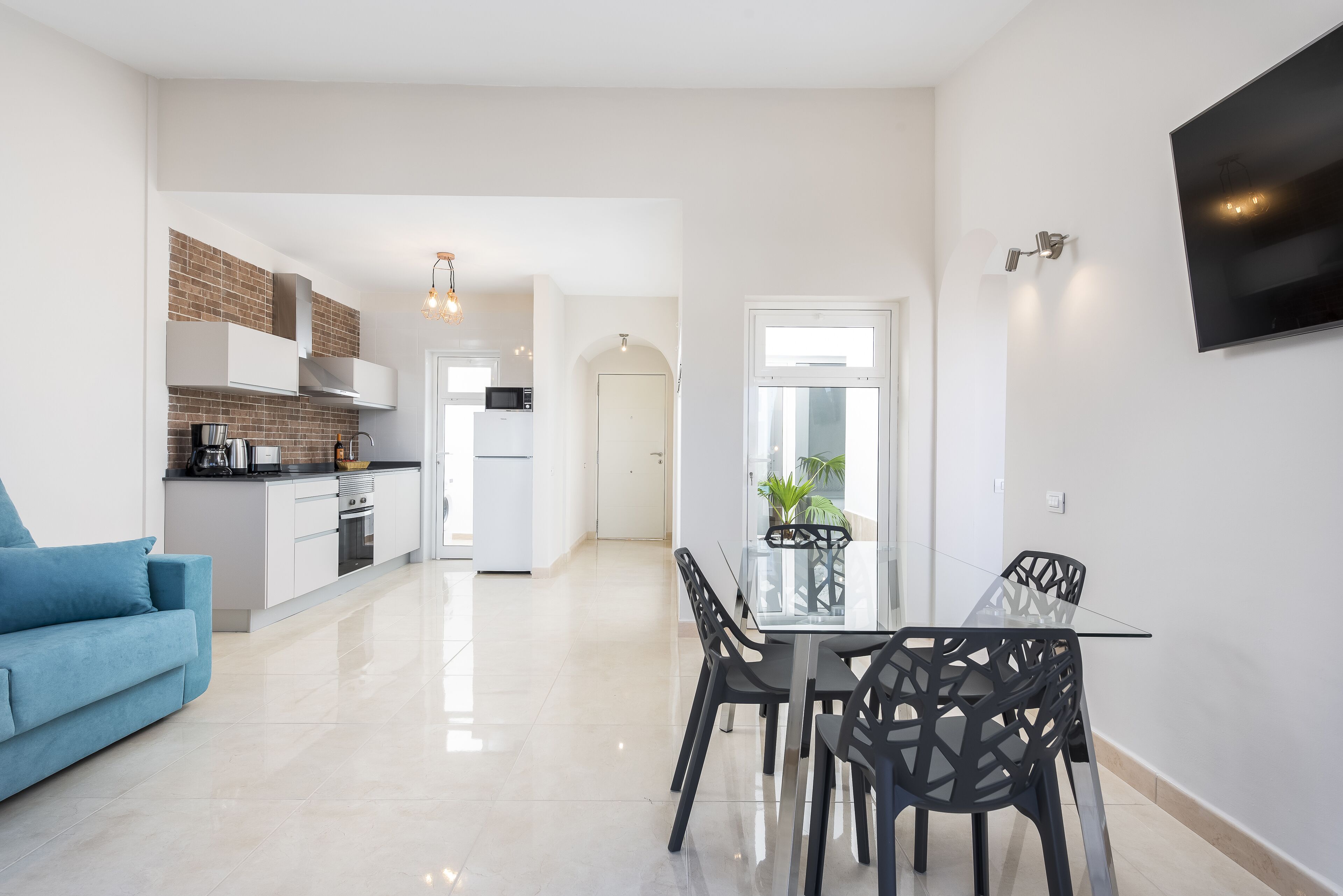 Una cocina moderna con armarios blancos, salpicadero de ladrillo y un comedor con mesa de vidrio y sillas negras.