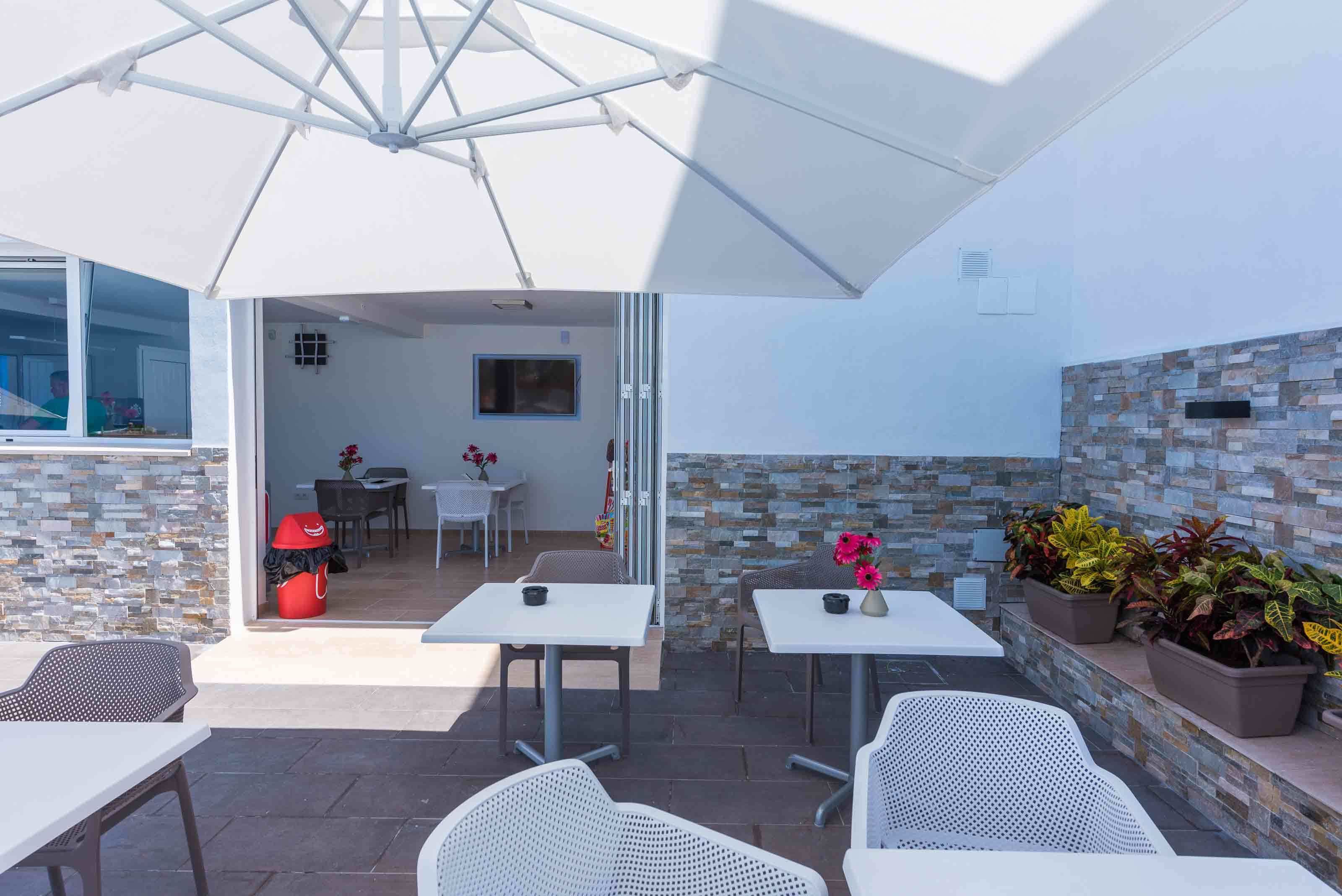 An outdoor patio area with white tables and chairs, a large umbrella, and potted plants.