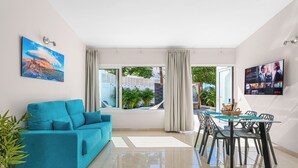A modern living room with a teal sofa, a glass dining table with chairs, a TV mounted on the wall, and a large window with curtains. - Haciendas Village Tenerife (Adeje)