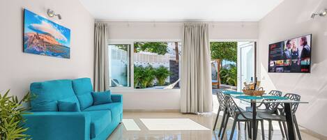 A modern living room with a teal sofa, a glass dining table with chairs, a TV mounted on the wall, and a large window with curtains.