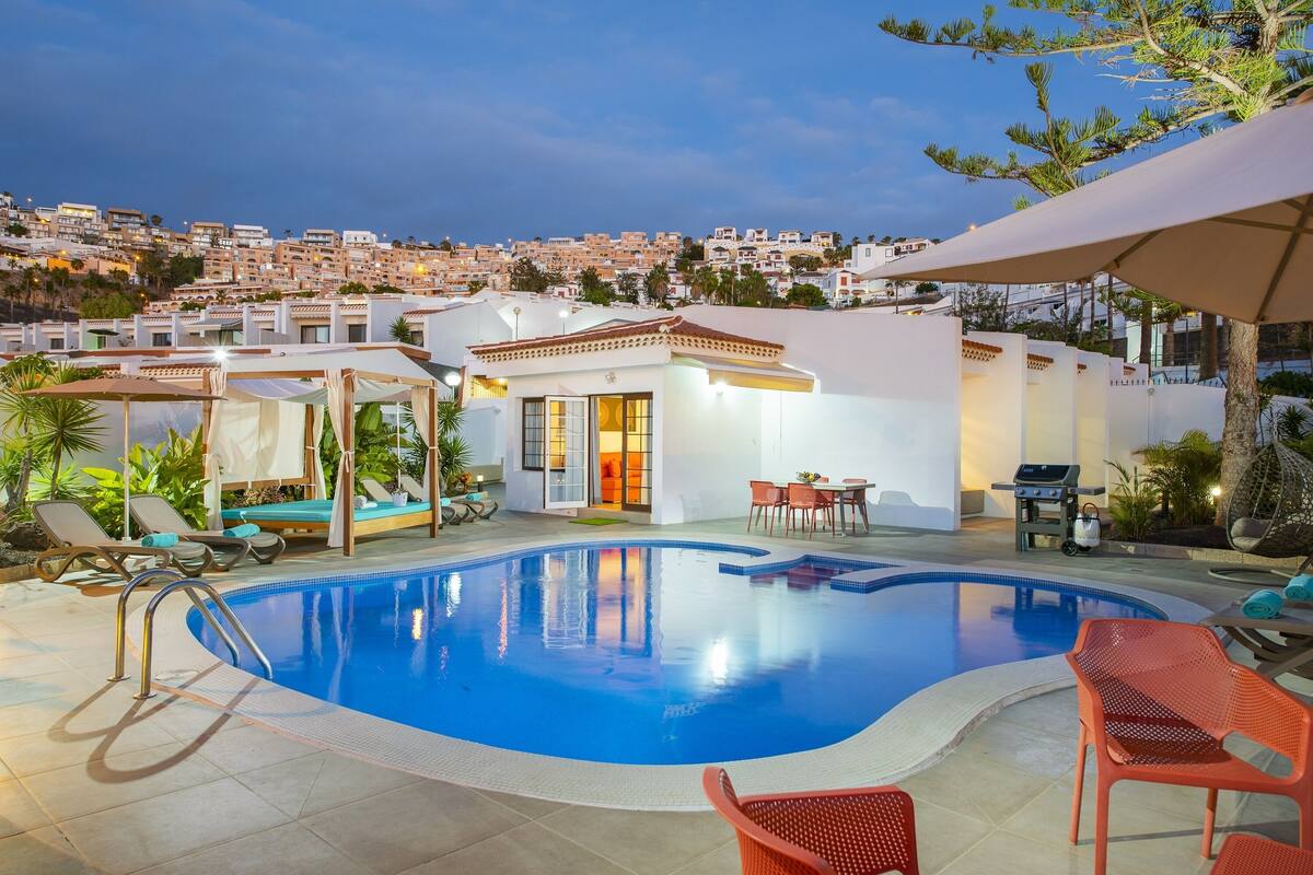 Una piscina con sillones y sombrillas, rodeada por un edificio blanco con vista a un pueblo en la ladera.