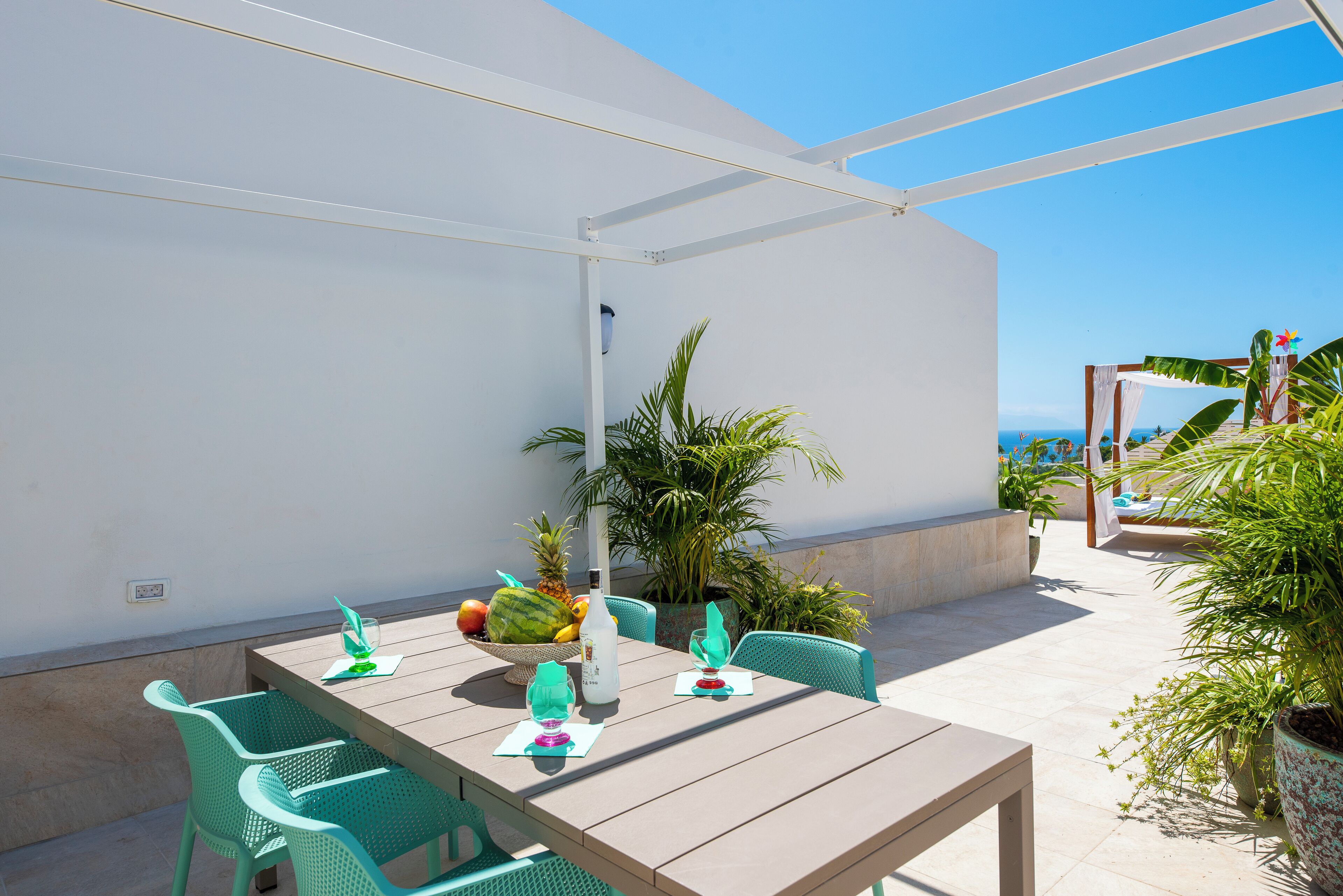 Un patio con mesa de madera y sillas verdes, rodeado de plantas en macetas y vistas al mar.