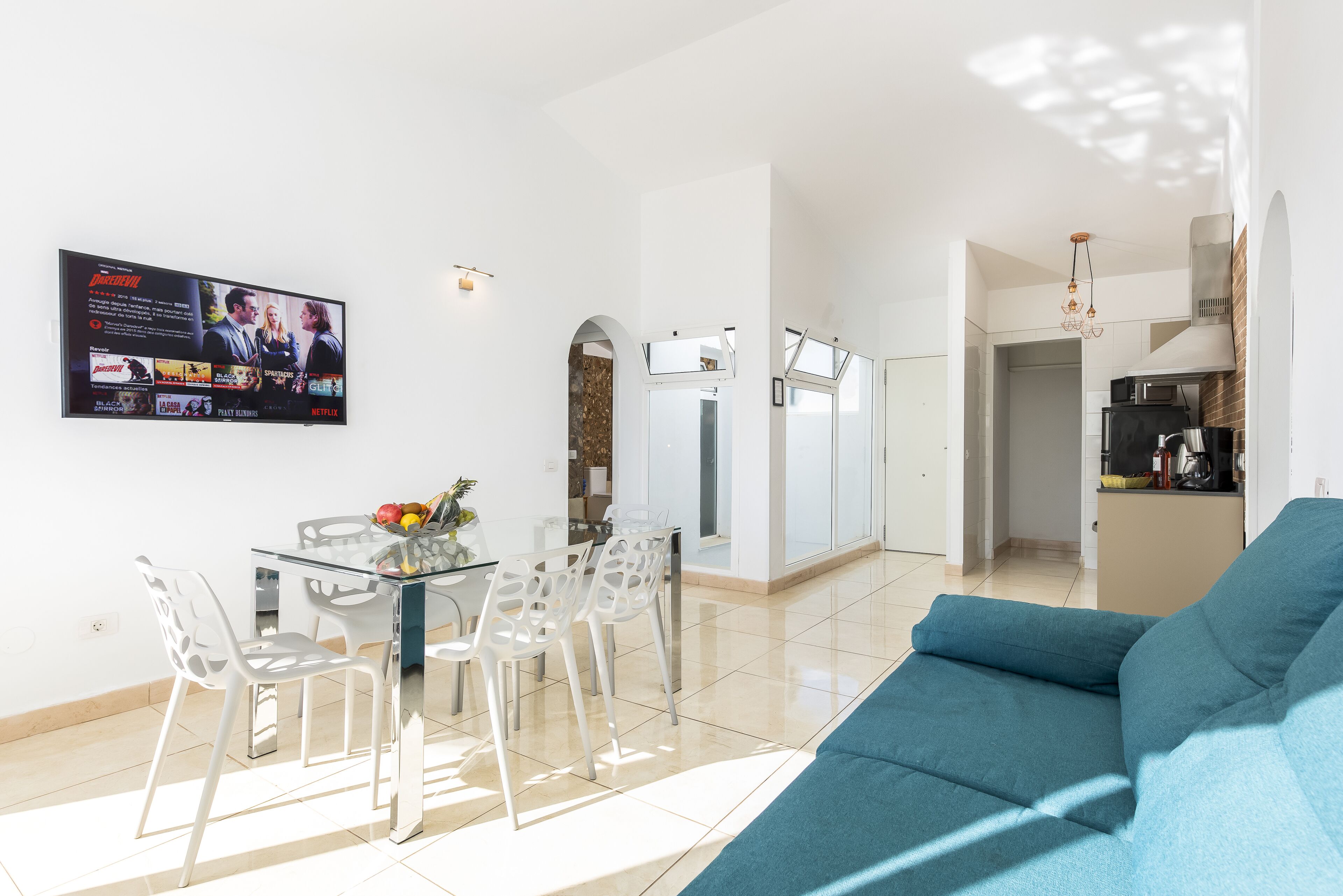 Una sala de estar moderna con un sofá azul, una mesa de comedor de vidrio con sillas, y un televisor de pantalla plana montado en la pared reproduciendo una película.