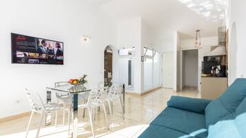 Una sala de estar moderna con un sofá azul, una mesa de comedor de vidrio con sillas, y un televisor de pantalla plana montado en la pared reproduciendo una película.
