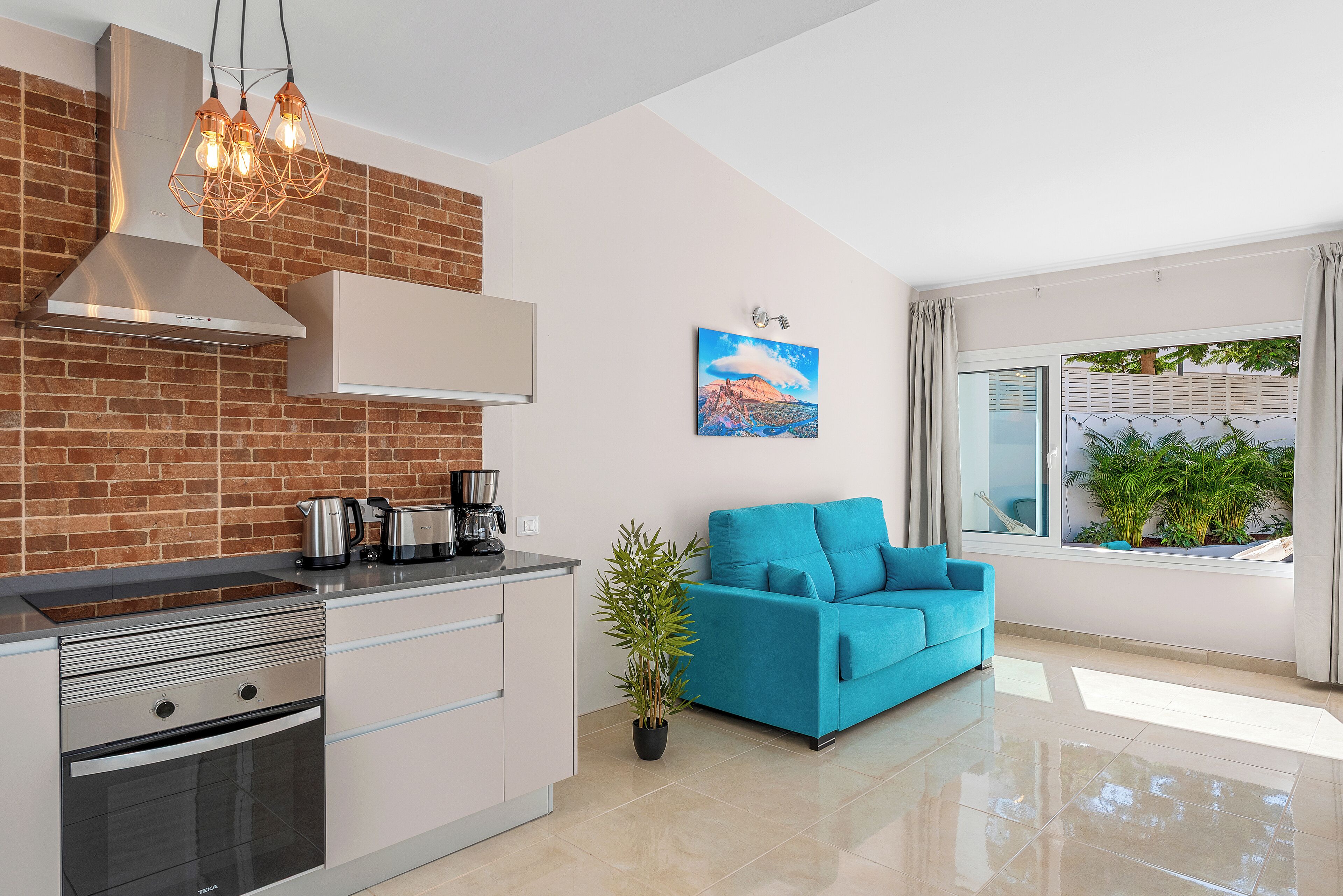 Une cuisine moderne avec un mur en briques, des appareils électroménagers en acier inoxydable et un canapé bleu placé près d’une fenêtre donnant sur un paysage verdoyant.