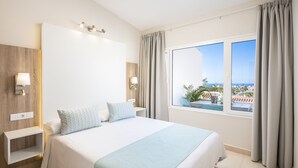 A hotel room with a large bed, a nightstand, a wall-mounted light, a window with curtains, and a view of the outside. - Haciendas Village Tenerife (Adeje)