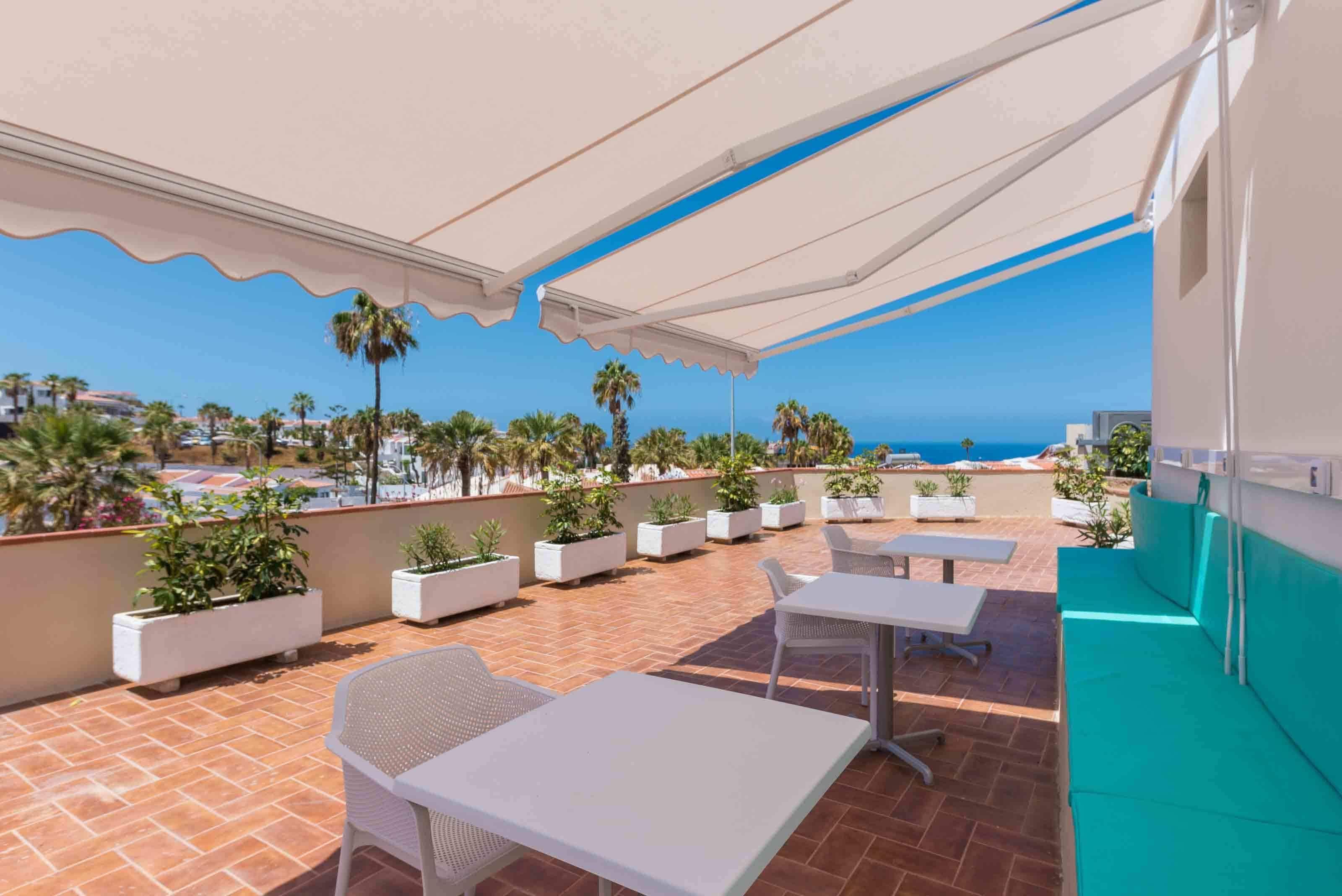 Un patio exterior con mesas y sillas blancas, plantas en macetas y vista al mar.