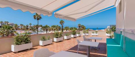 Un patio exterior con mesas y sillas blancas, plantas en macetas y vista al mar.