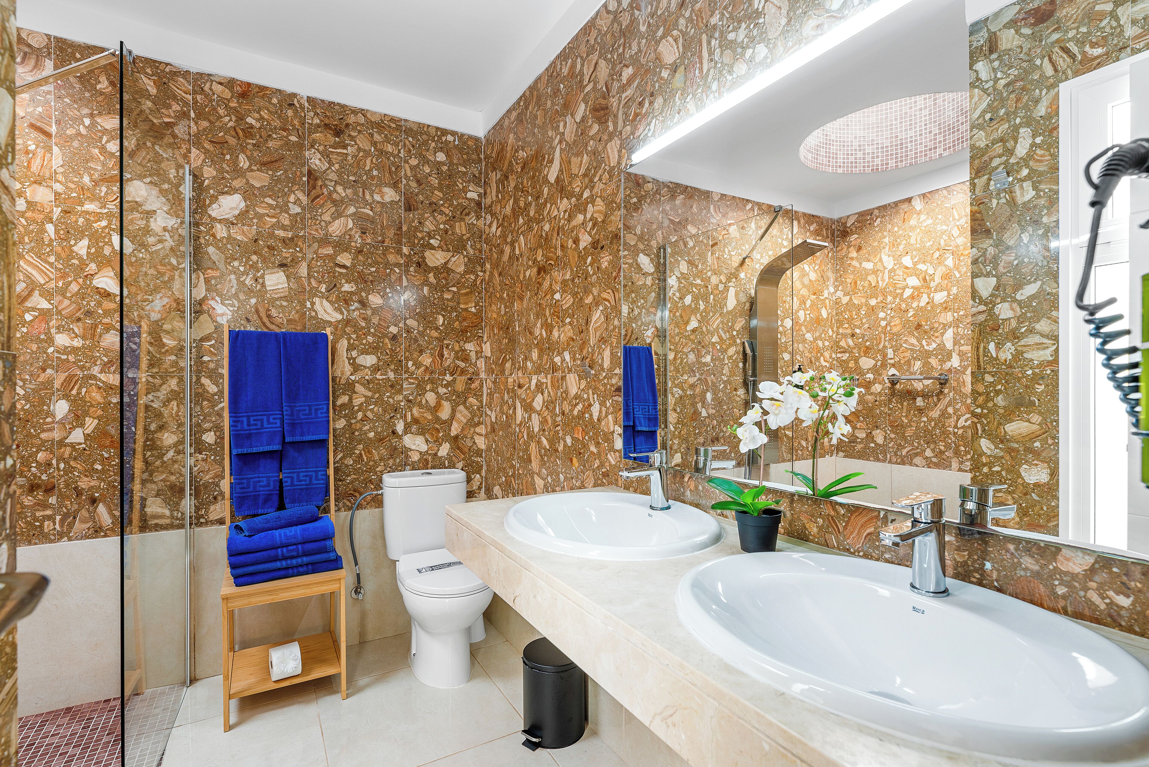 Une salle de bain avec des murs en marbre, deux lavabos, des toilettes et une douche à parois de verre.