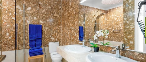 Une salle de bain avec des murs en marbre, deux lavabos, des toilettes et une douche à parois de verre.