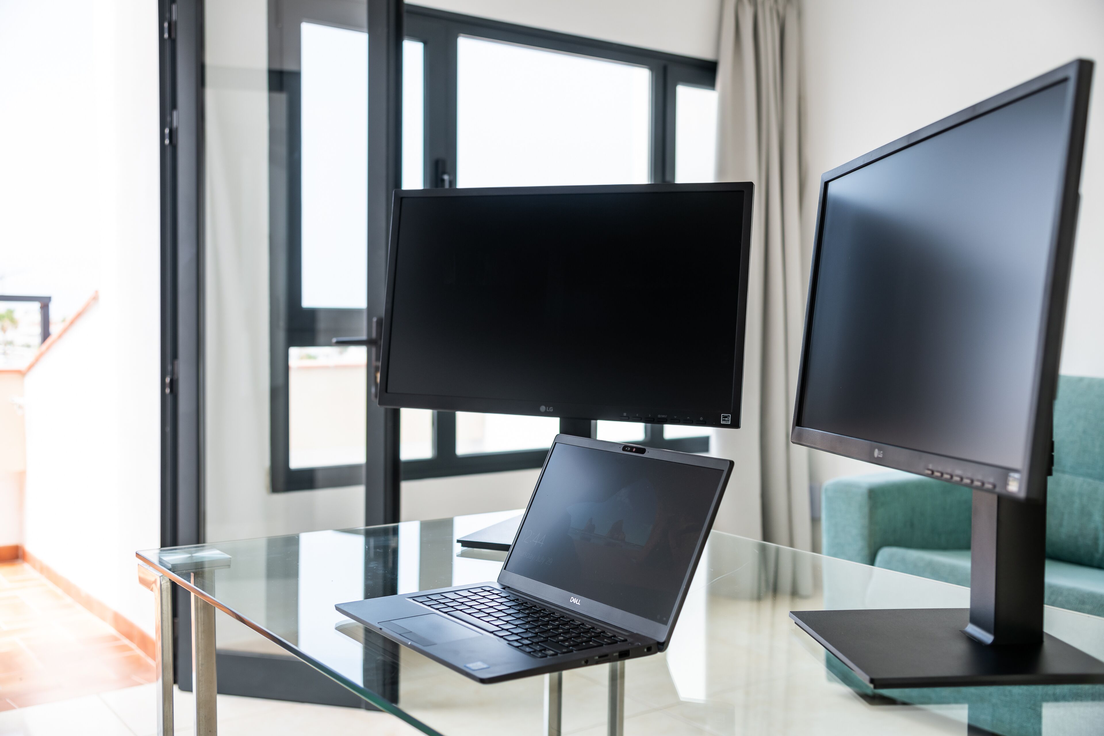 A modern room with a glass table holding a laptop and two monitors, a teal sofa, and a large window with curtains.