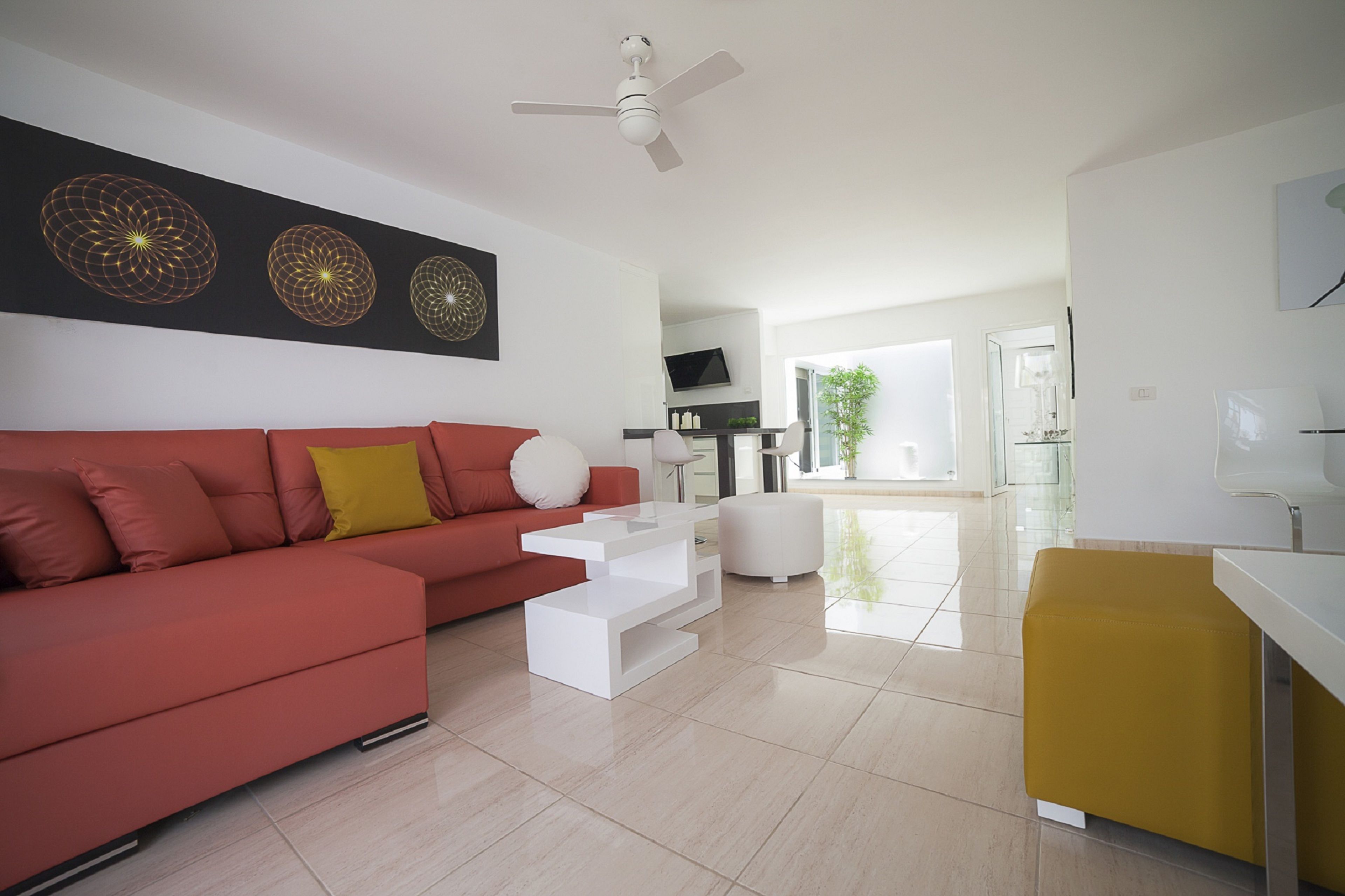 A modern living room with a red sofa, white coffee table, and a yellow ottoman. There is a ceiling fan, a flat-screen TV, and a kitchen area in the background.