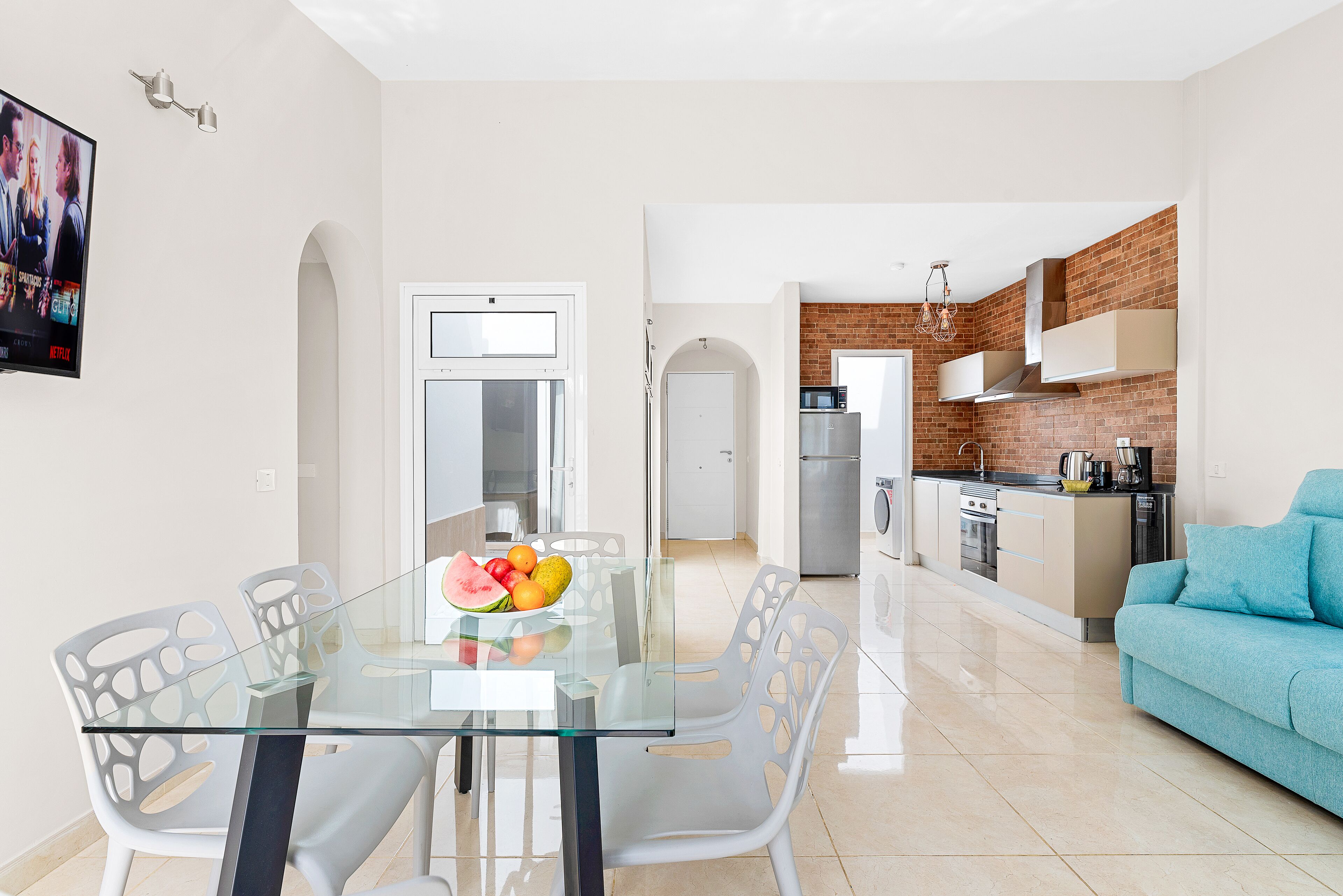 A modern kitchen with a brick backsplash, a dining area with a glass table and white chairs, and a blue sofa in the living room.