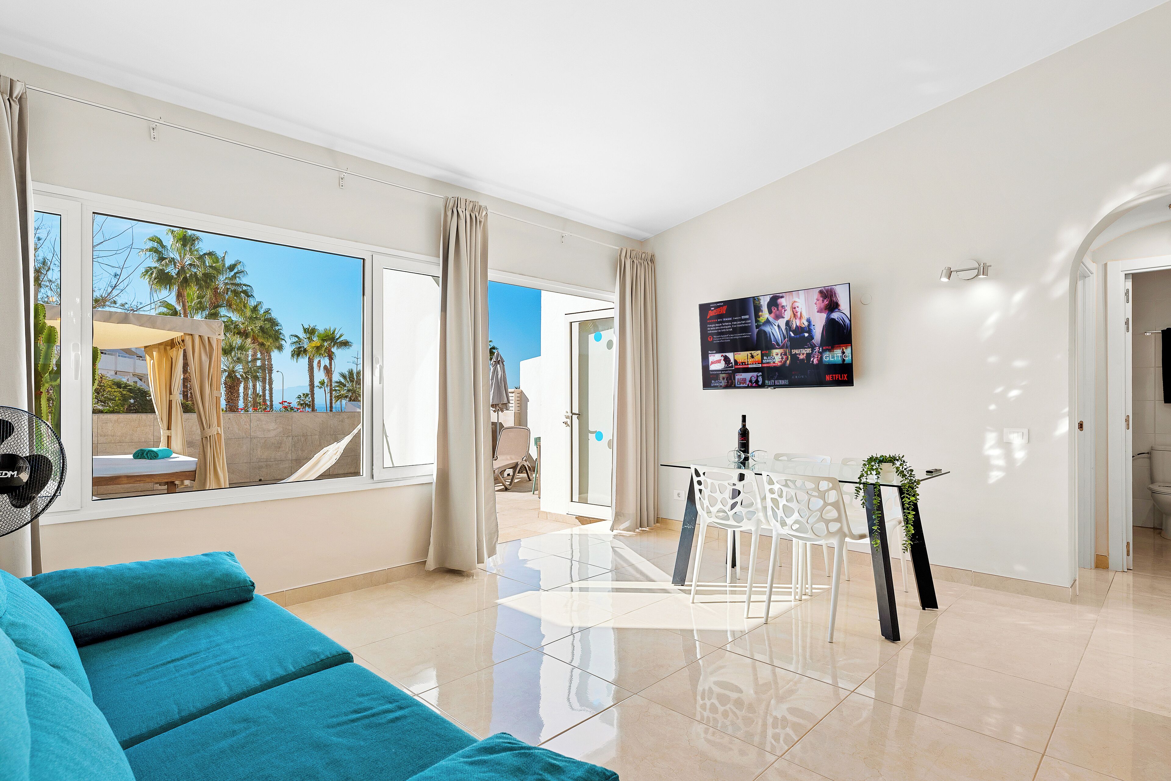 Una sala de estar moderna con un sofá, una mesa de comedor con sillas, un televisor montado en la pared y ventanas grandes con cortinas.