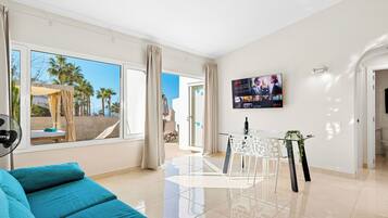 Una sala de estar moderna con un sofá, una mesa de comedor con sillas, un televisor montado en la pared y ventanas grandes con cortinas.