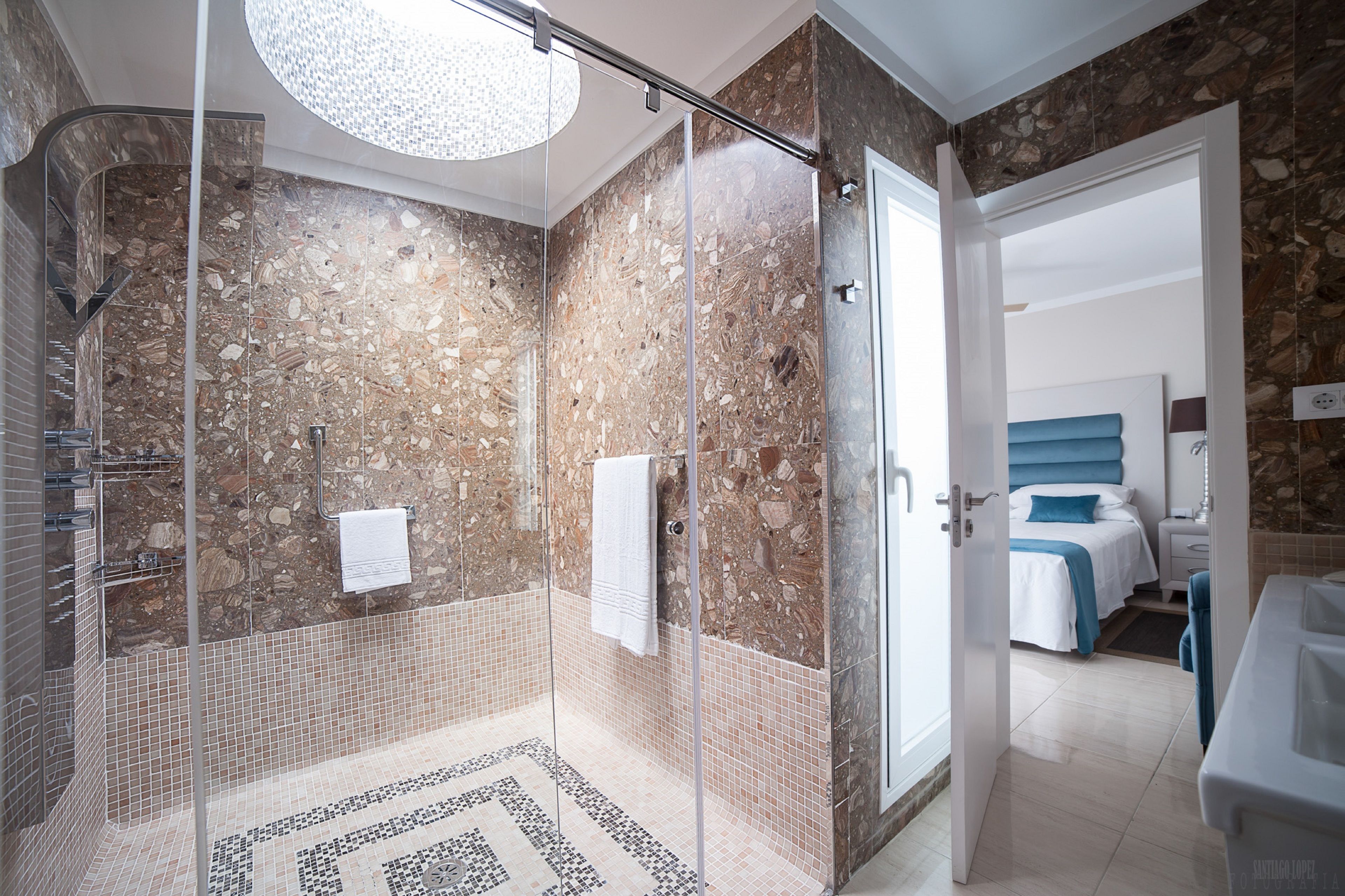 Une salle de bain moderne dotée d’une douche en verre, d’un mur en marbre et d’une chambre à coucher visible, avec un lit et une table de chevet.