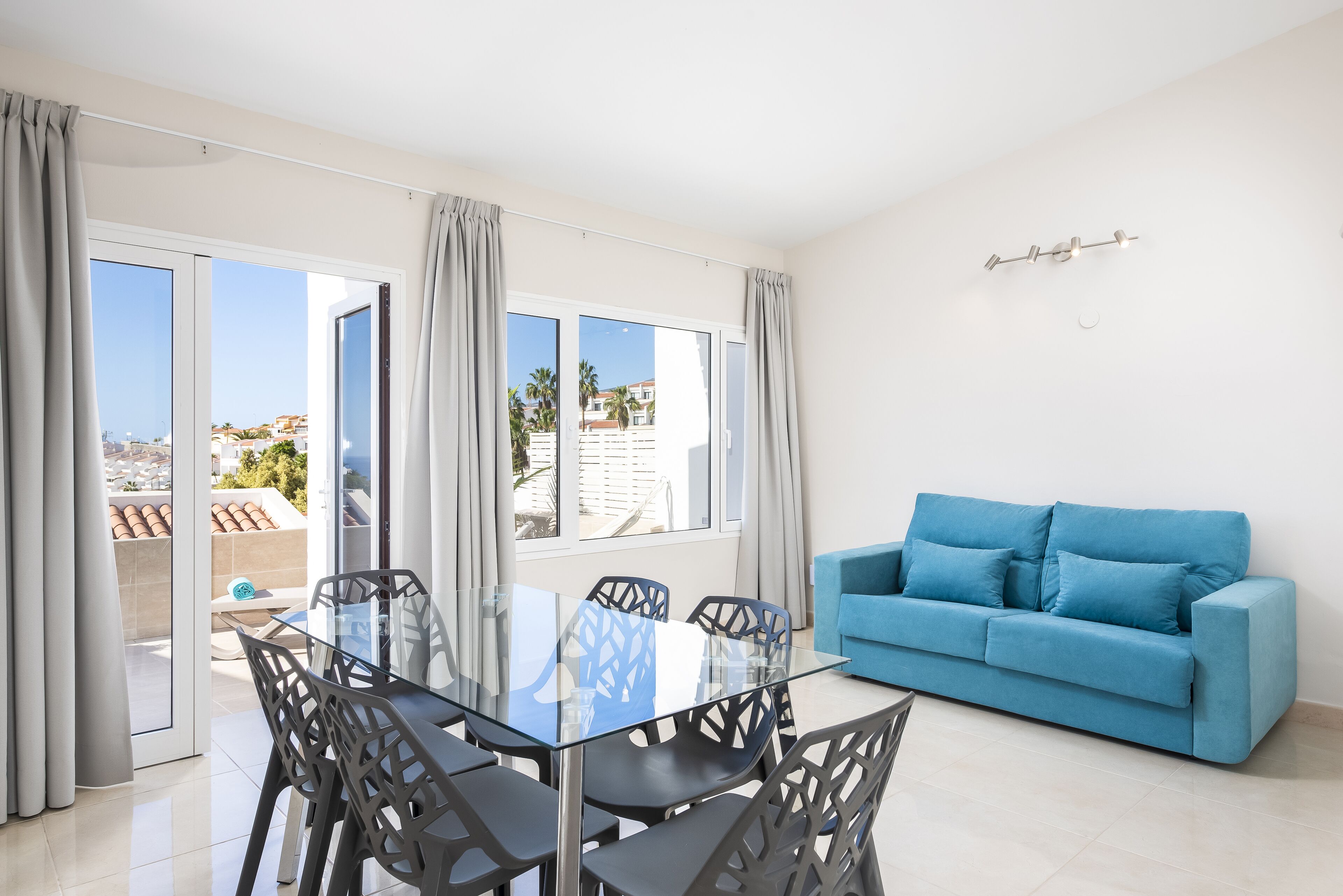 A modern living room with a glass dining table, blue sofa, and a balcony with curtains.