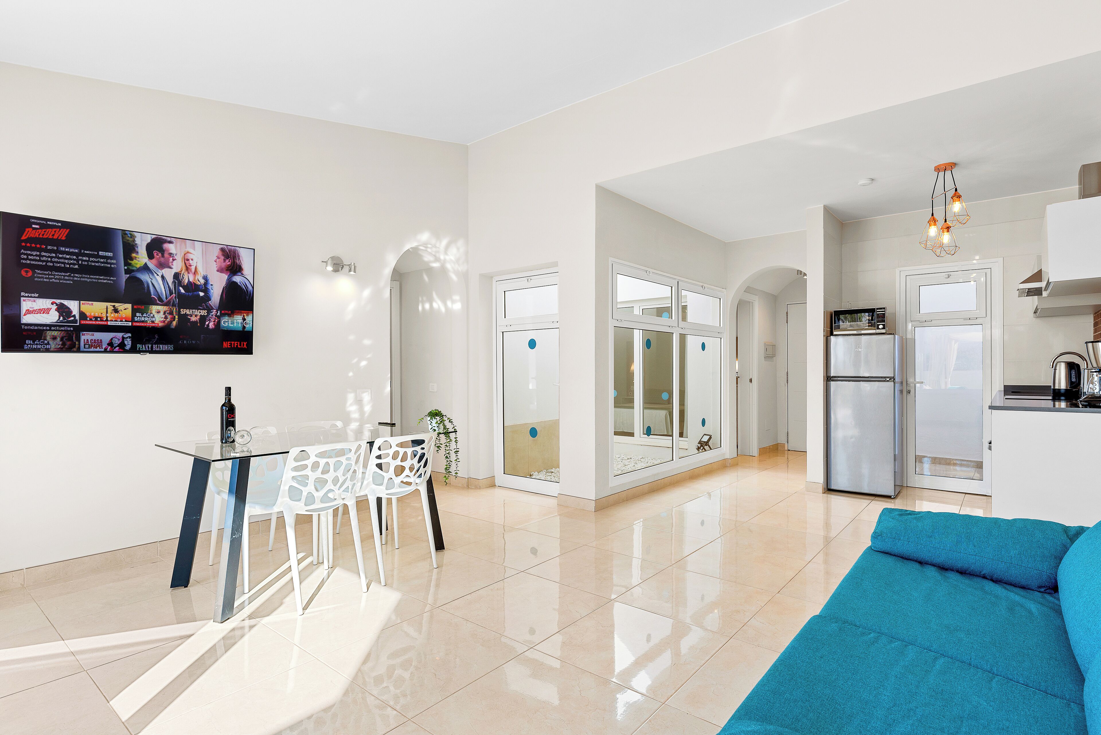 A modern living room with a glass dining table, white chairs, a blue sofa, and a kitchen area with a refrigerator and a TV displaying a movie poster.