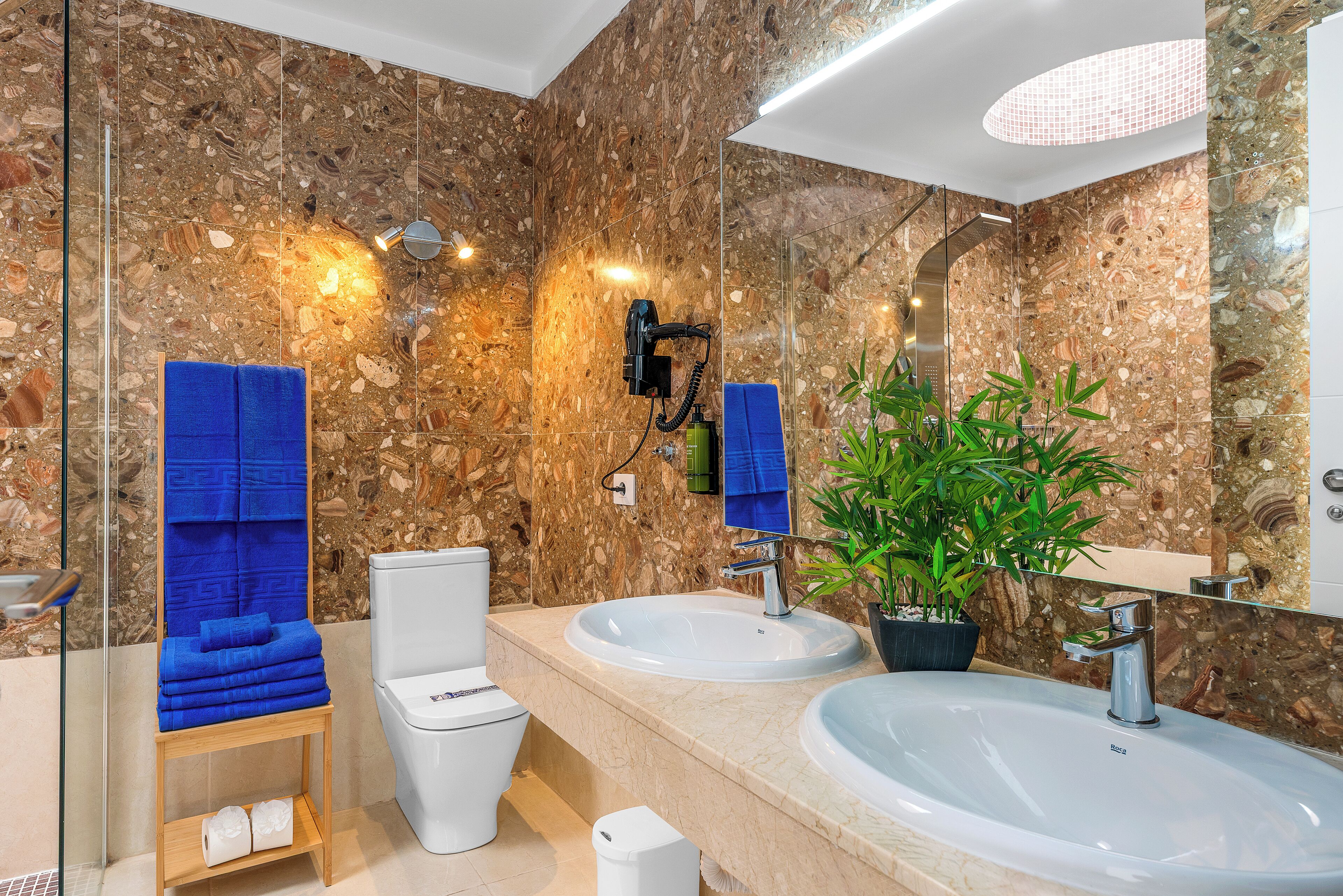 A modern bathroom with a marble backsplash, a large mirror, two sinks, a toilet, and a blue towel rack.