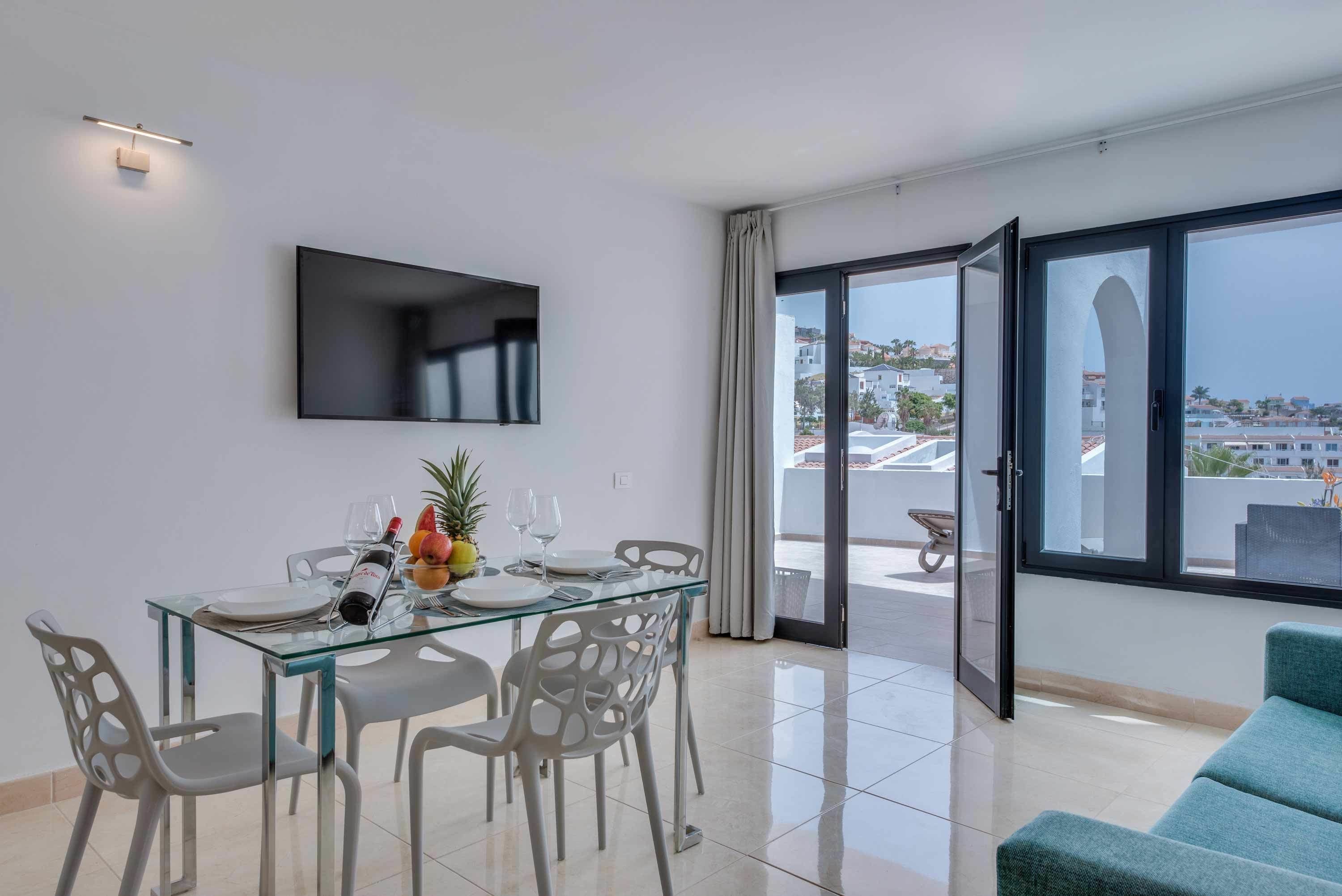 Un soggiorno moderno con tavolo da pranzo in vetro, sedie bianche, TV a schermo piatto montata a parete e un'ampia porta scorrevole in vetro che dà su un balcone esterno.