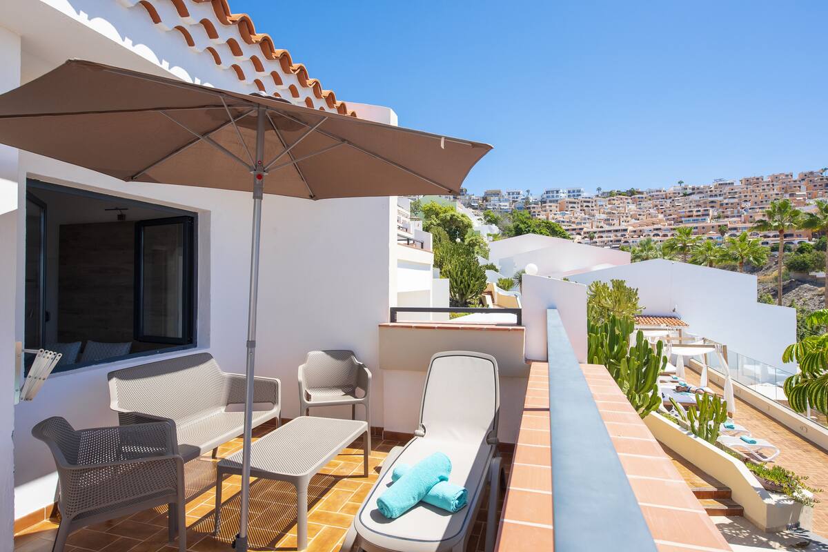 Terraza en la azotea con zona de estar, que incluye una mesa, sillas y una tumbona, con vistas a una zona residencial densamente poblada.