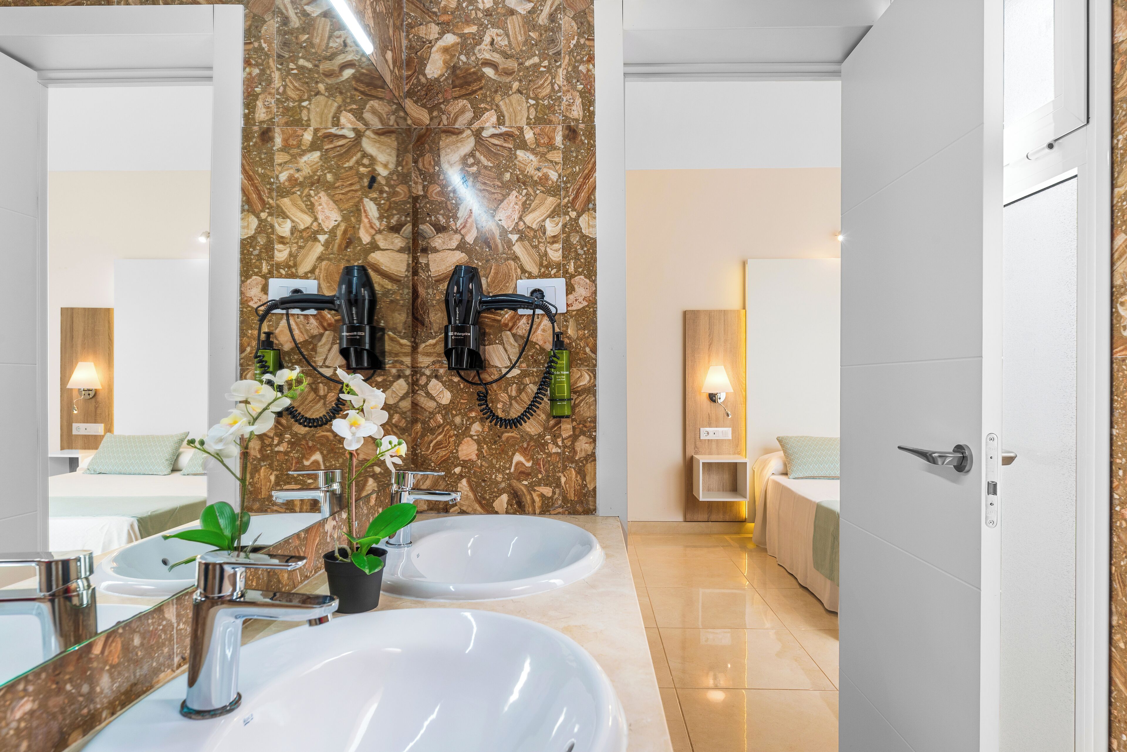 A modern bathroom with two sinks, a marble backsplash, and a bed visible through an open door.