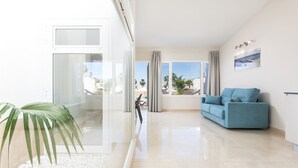A modern living room with a blue sofa, a glass partition, and a view of palm trees and buildings outside. - Haciendas Village Tenerife (Adeje)