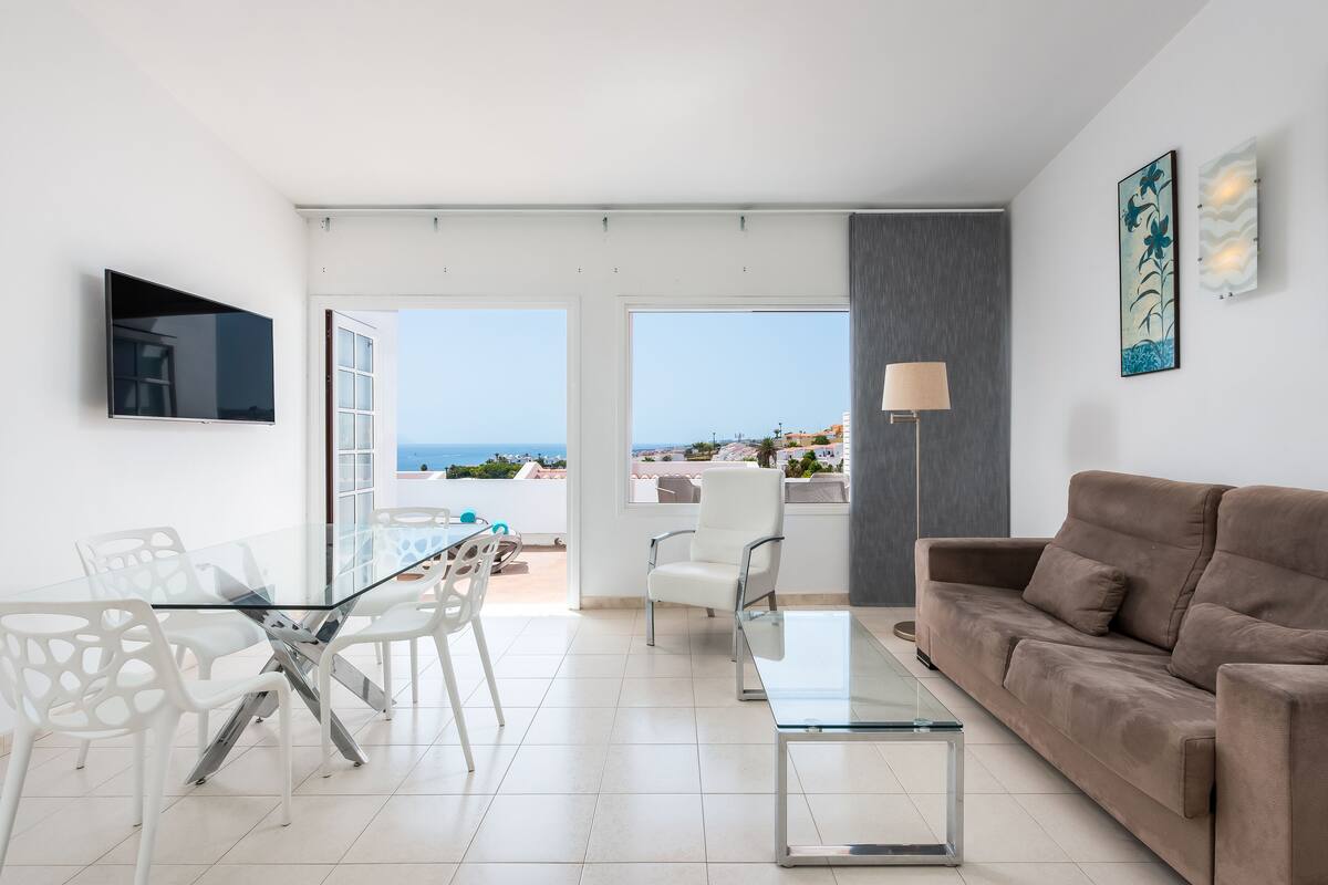 Uma sala de estar moderna com uma mesa e cadeiras de vidro, um sofá, uma TV e uma varanda com vista para o mar.