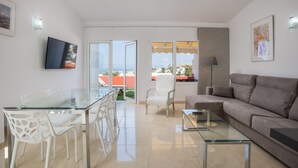 Ein modernes Wohnzimmer mit einem Glastisch, weißen Stühlen, einem grauen Sofa und einem an der Wand befestigten Fernseher.