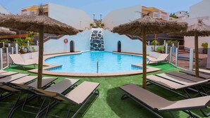 Ein Pool mit Liegestühlen und Reetdachsonnenmarkisen, umgeben von viel Grün und weißen Gebäuden.