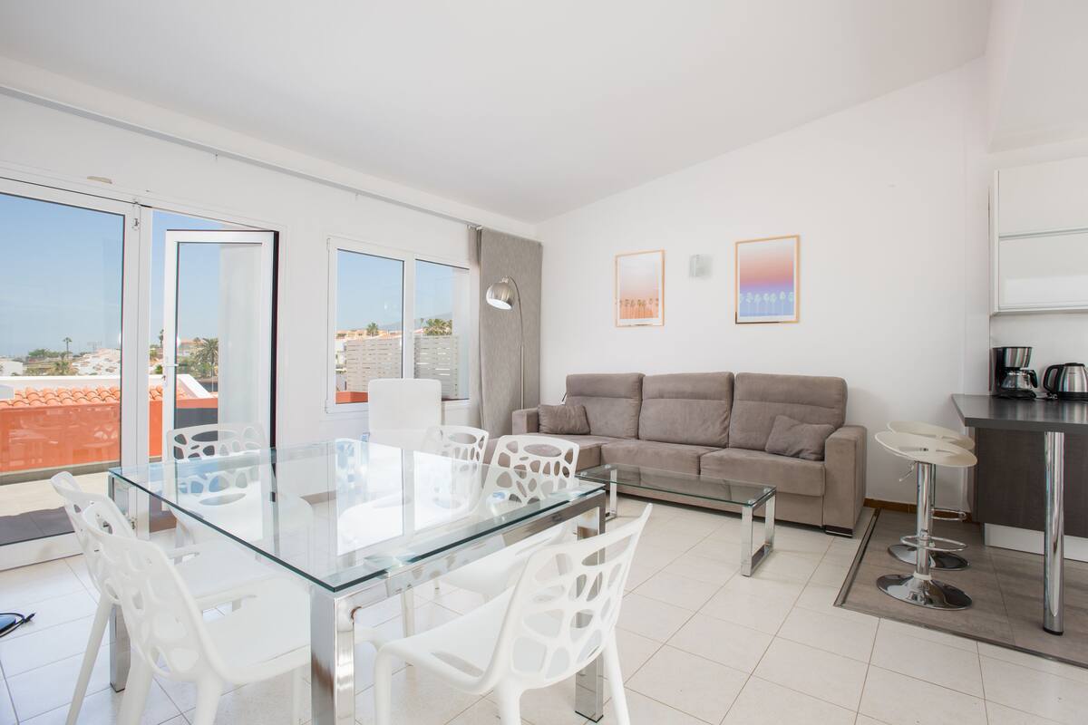 Uma sala moderna com uma mesa de jantar de vidro, cadeiras brancas, um sofá e uma área de cozinha.