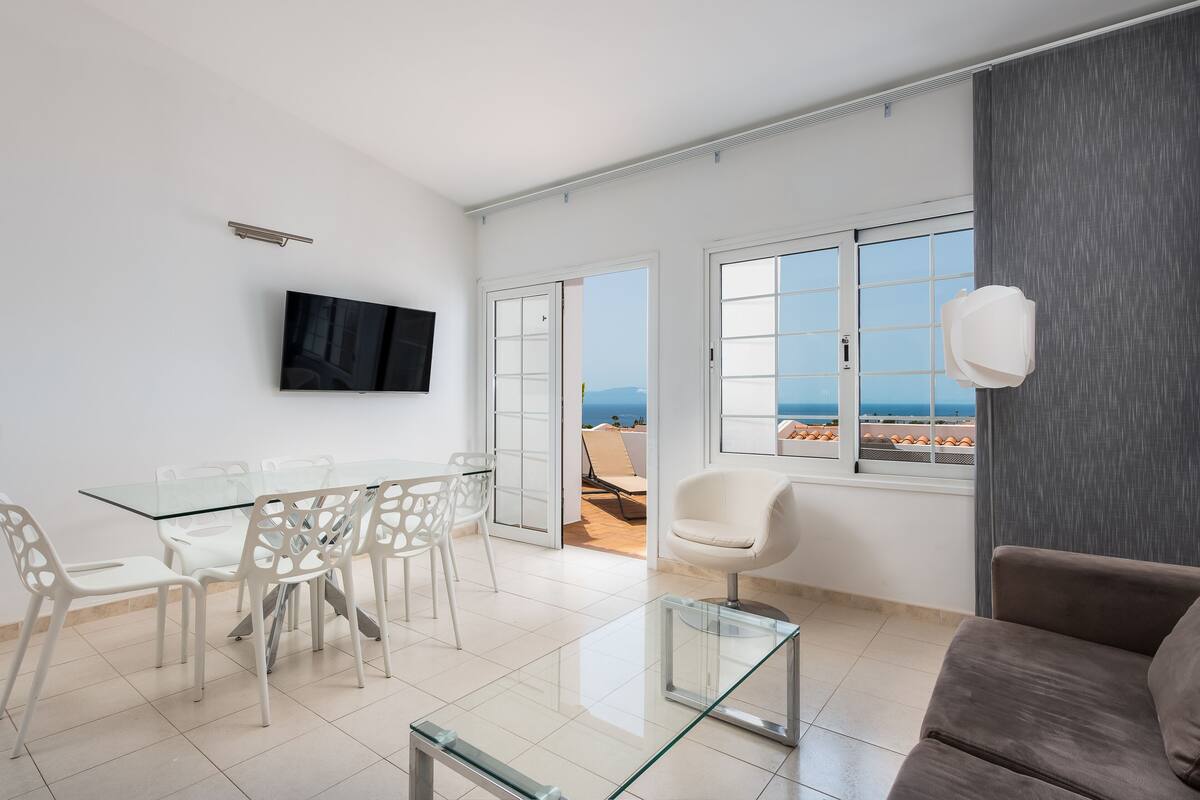 Uma sala de estar moderna com uma mesa de jantar de vidro, cadeiras brancas, um sofá marrom e uma TV fixada na parede.