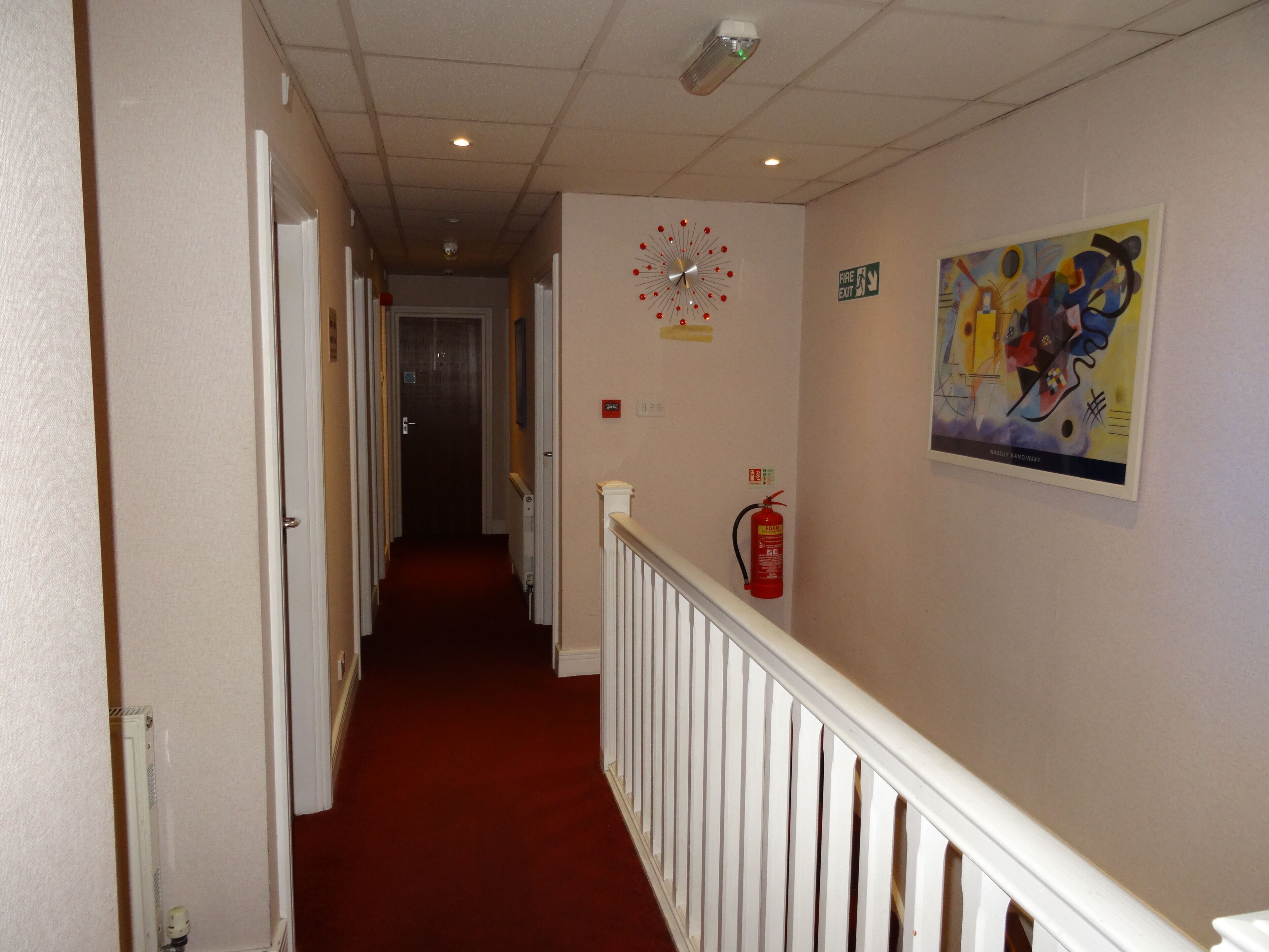 A hallway with a red carpet, white walls, a clock, a fire exit sign, and a fire extinguisher.