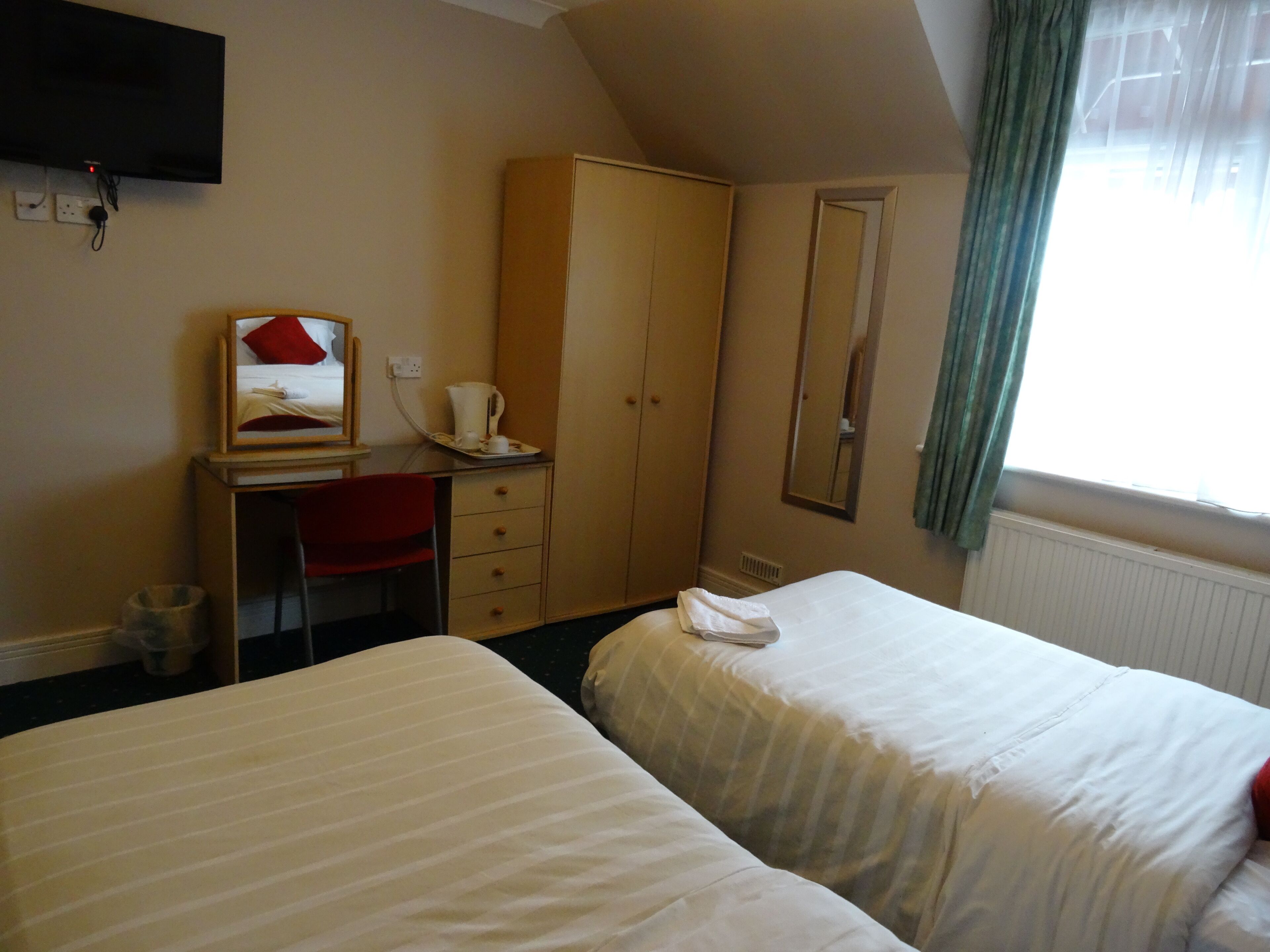 A hotel room with two beds, a desk, a chair, a wardrobe, a mirror, and a window with curtains.