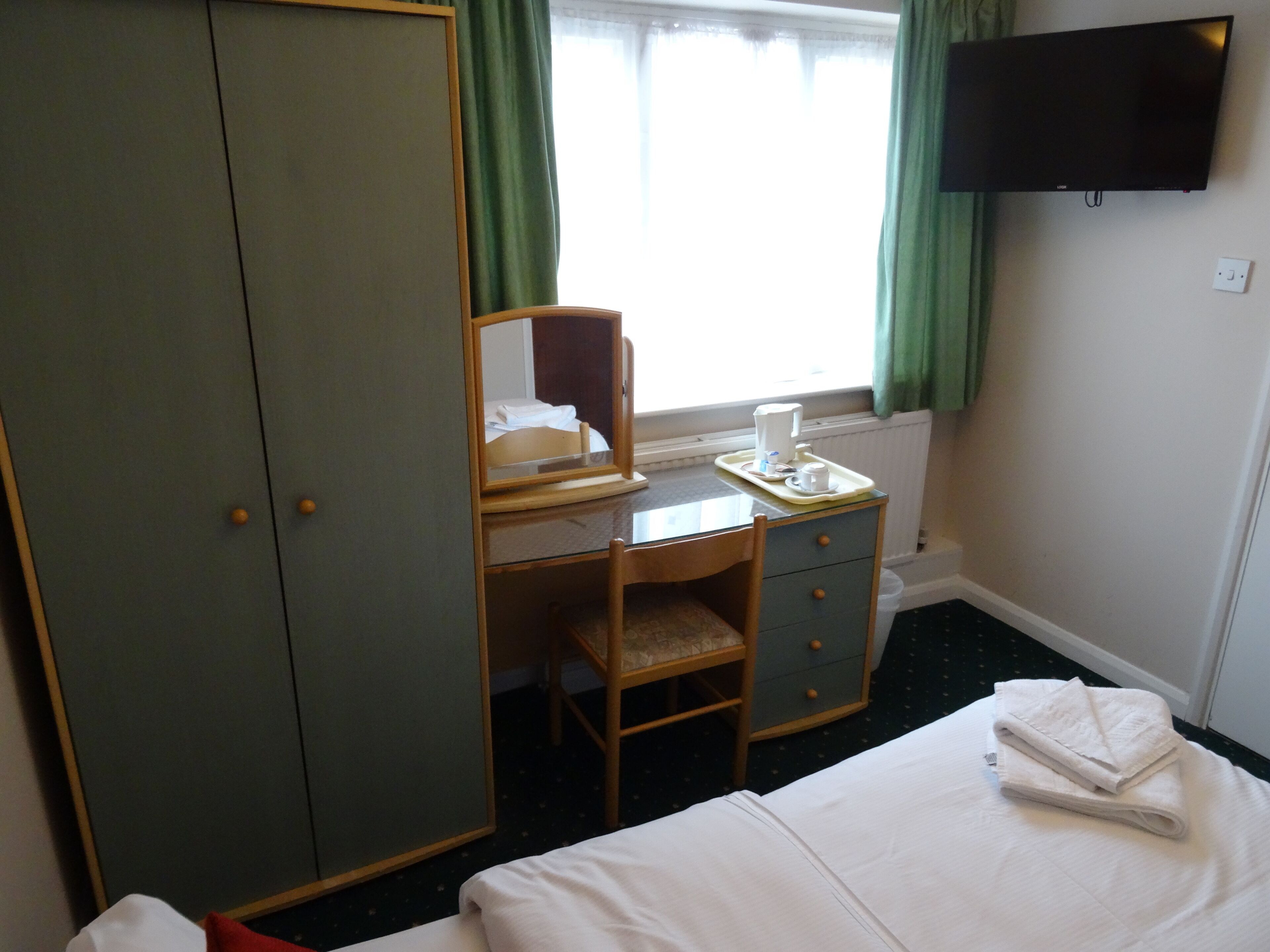 A hotel room with a bed, a desk with a chair, a wardrobe, a mirror, a window with curtains, and a flat-screen TV mounted on the wall.