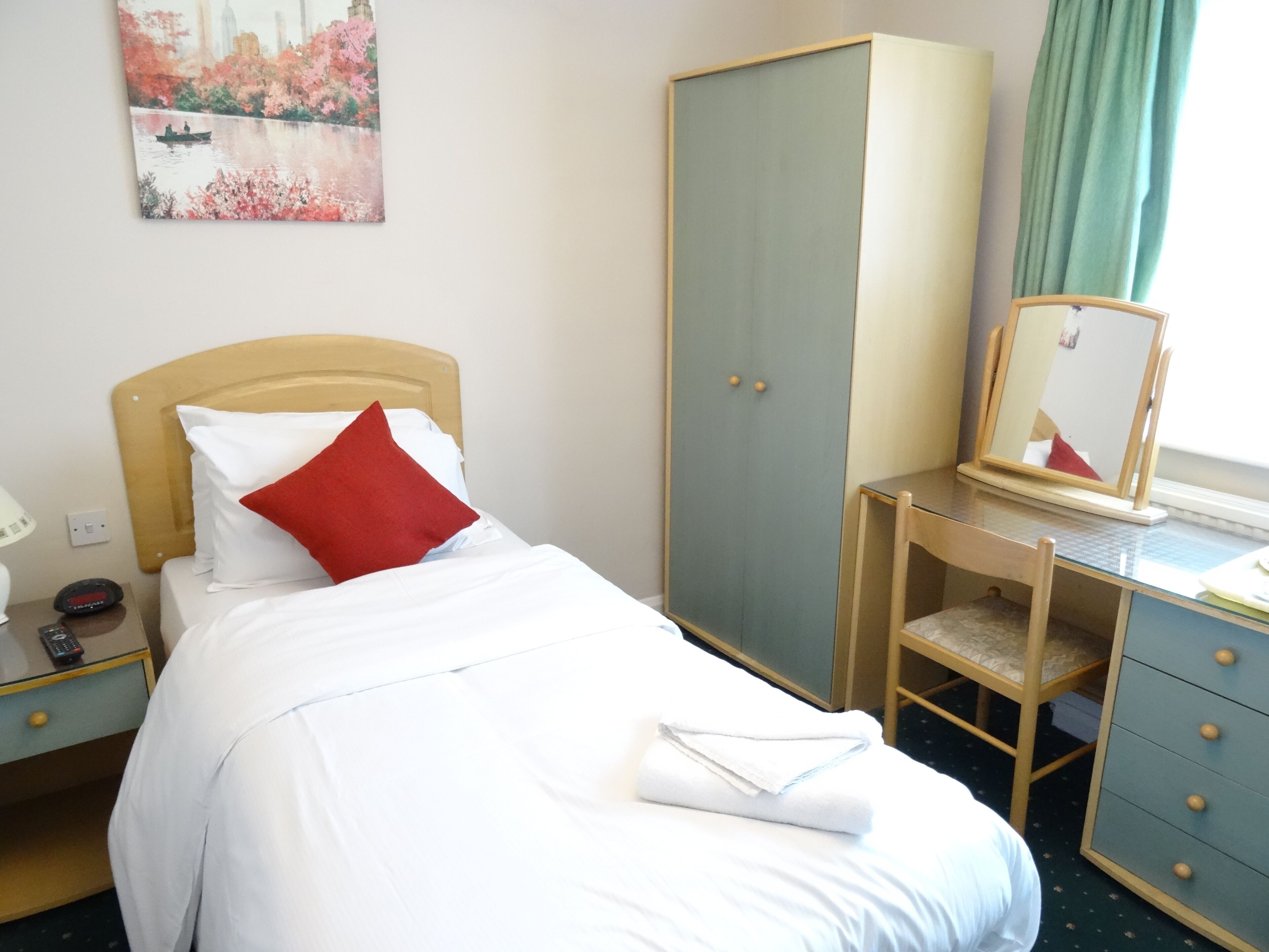 A hotel room with a bed, a wardrobe, a desk with a chair, a bedside table with a lamp, and a painting on the wall.