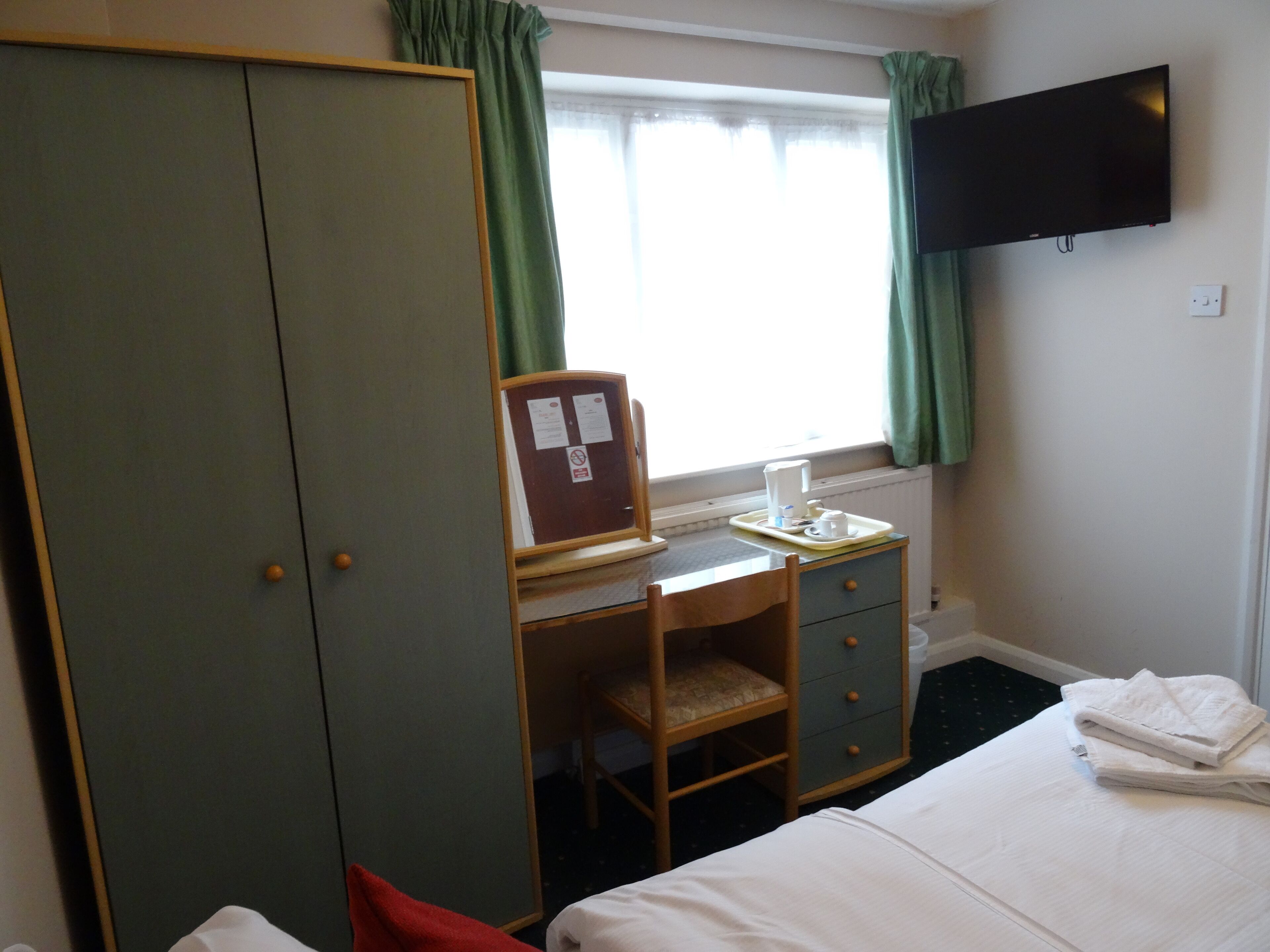 Habitación de hotel con cama, escritorio y silla, armario, televisor en la pared y ventana con cortinas.