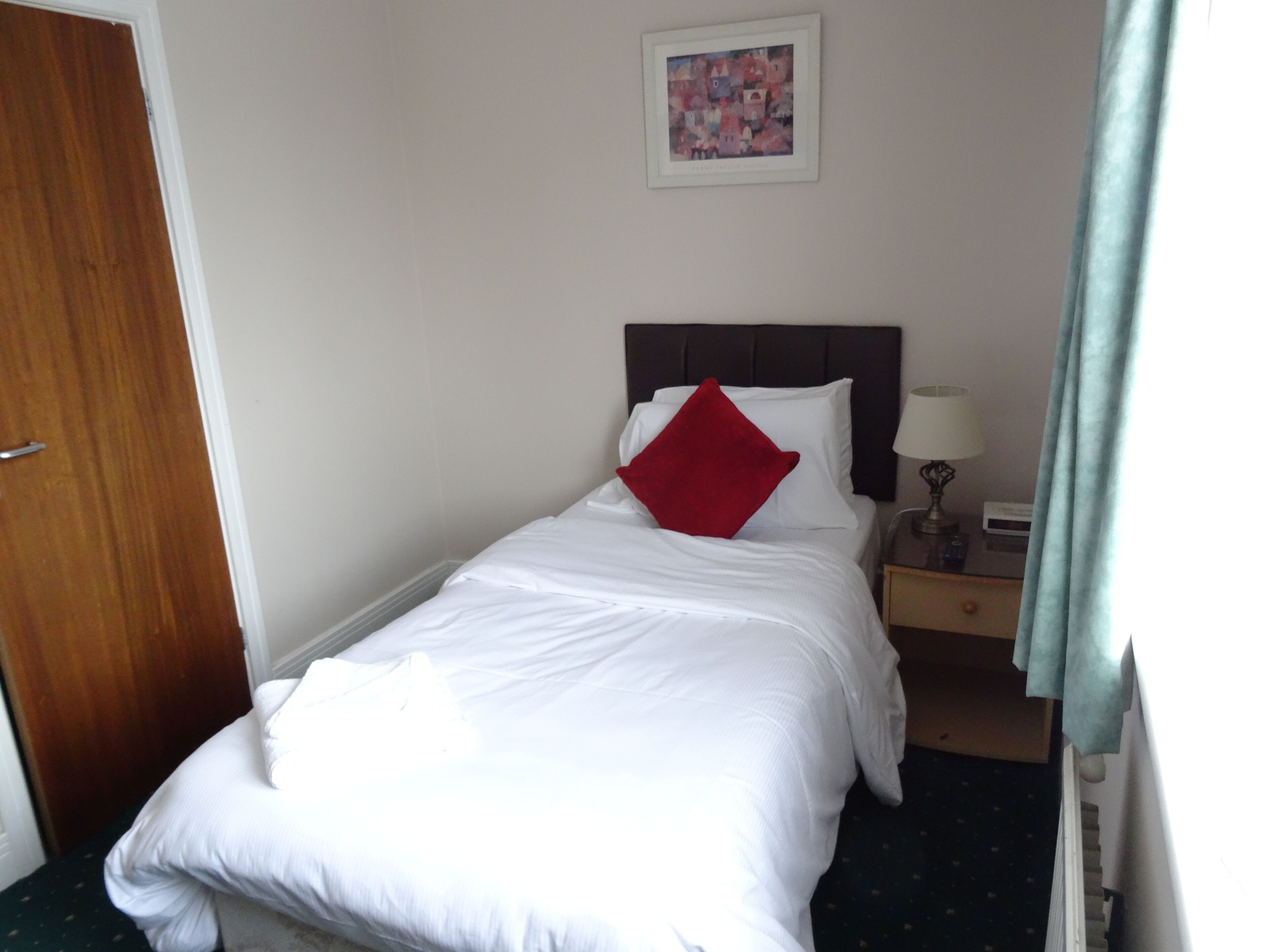 A neatly made bed with white linens and a red pillow, a bedside table with a lamp, and a framed picture on the wall.