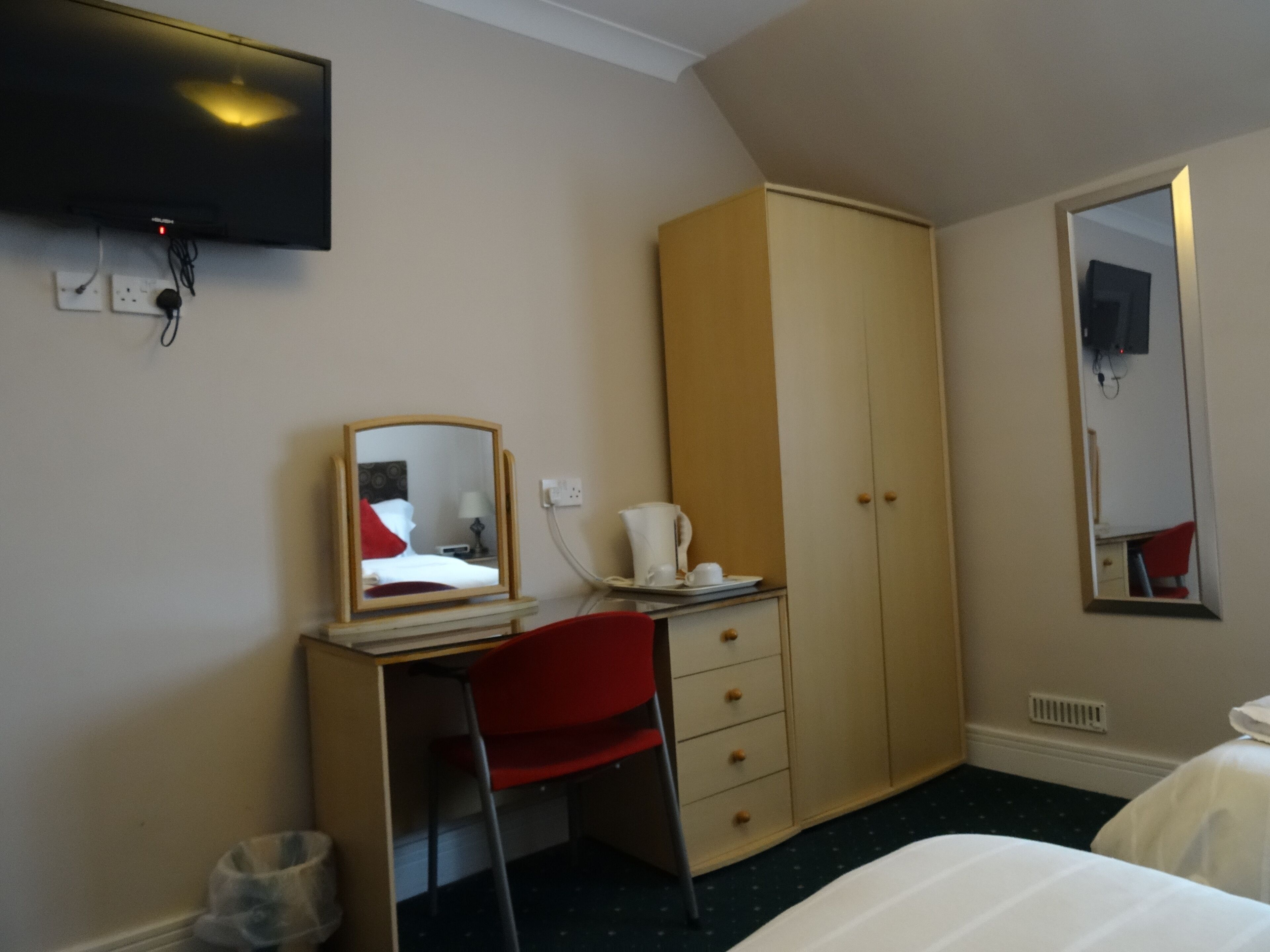 A hotel room with a desk, a red chair, a wardrobe, a bed, and a TV mounted on the wall.