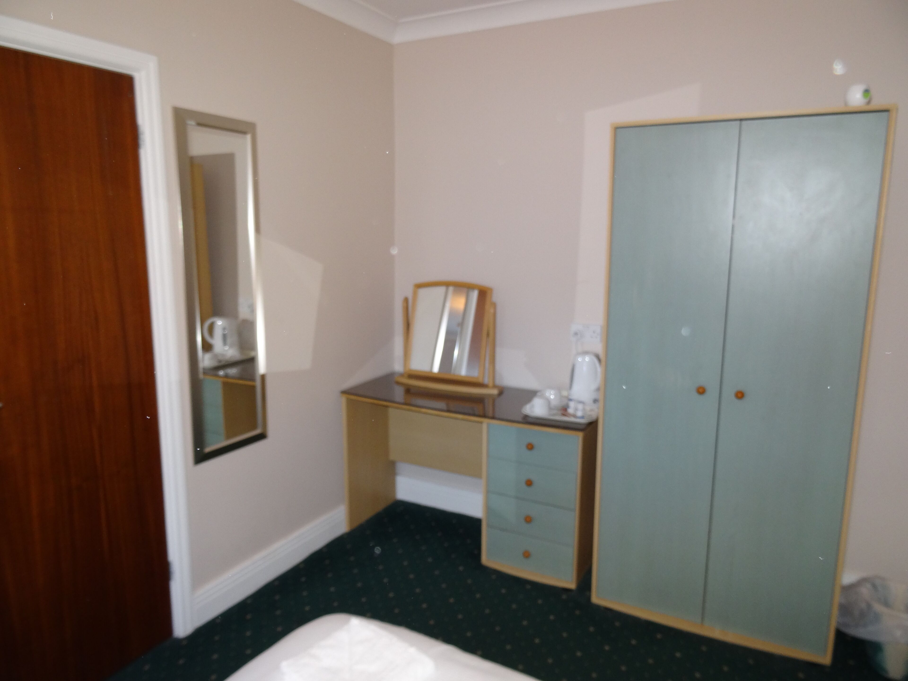 Une petite pièce simple, avec une porte en bois, un bureau doté d’un miroir, une armoire et un lavabo blanc.