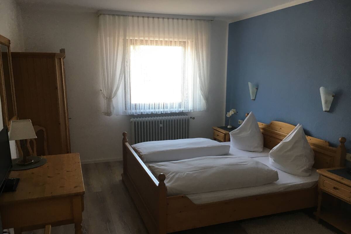 A bedroom with a wooden bed, a desk with a lamp, a radiator, and a window with sheer curtains.