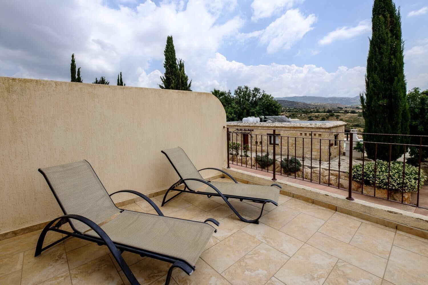 A balcony with two lounge chairs, a stone wall, and a view of a distant landscape with trees and buildings.