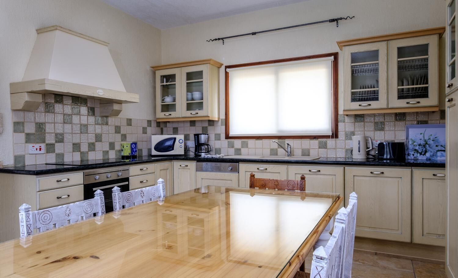 Una cocina con mesa de madera, armarios blancos, microondas y ventana con persianas.