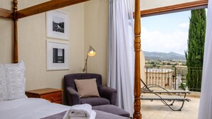 A bedroom with a four-poster bed, a chair, a nightstand, and a balcony with a view of the countryside. - Villa Anemona (Pegeia)