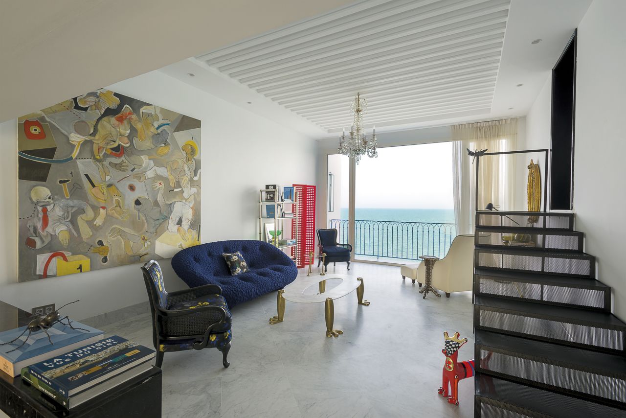 Un soggiorno spazioso con un'ampia pittura astratta, un divano blu, un tavolino da caffè e una scala che conduce a un balcone con vista sul mare.